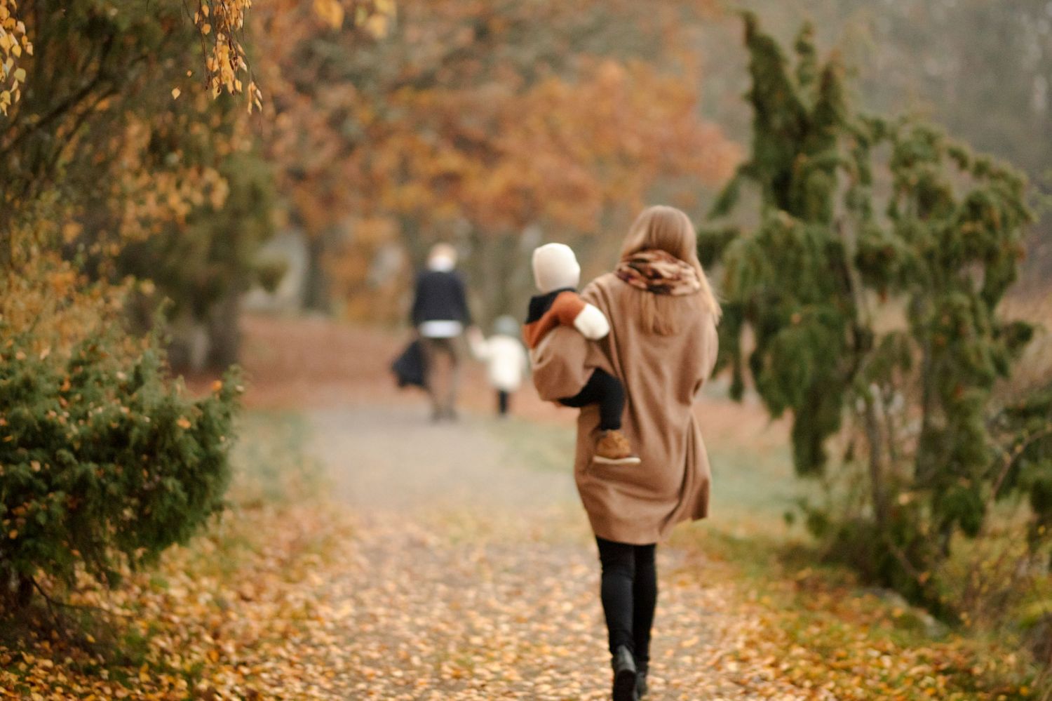 Someone in a brown coat walks along an autumn path covered in fallen leaves.