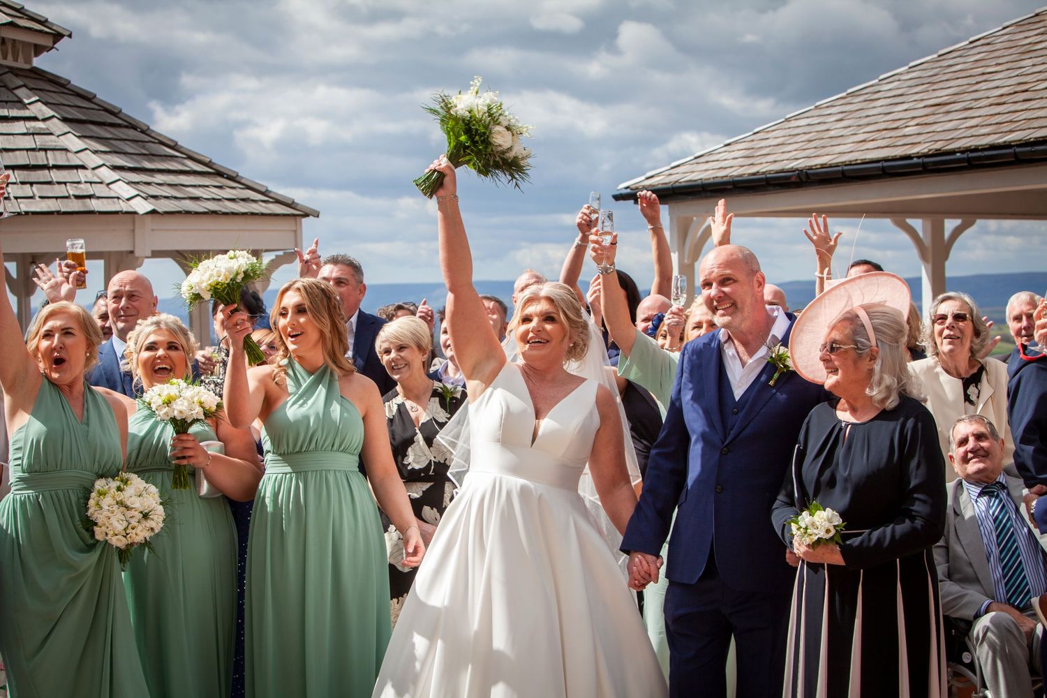 Joyful bride and groom celebrate with raised hands surrounded by cheering guests and bridesmaids.