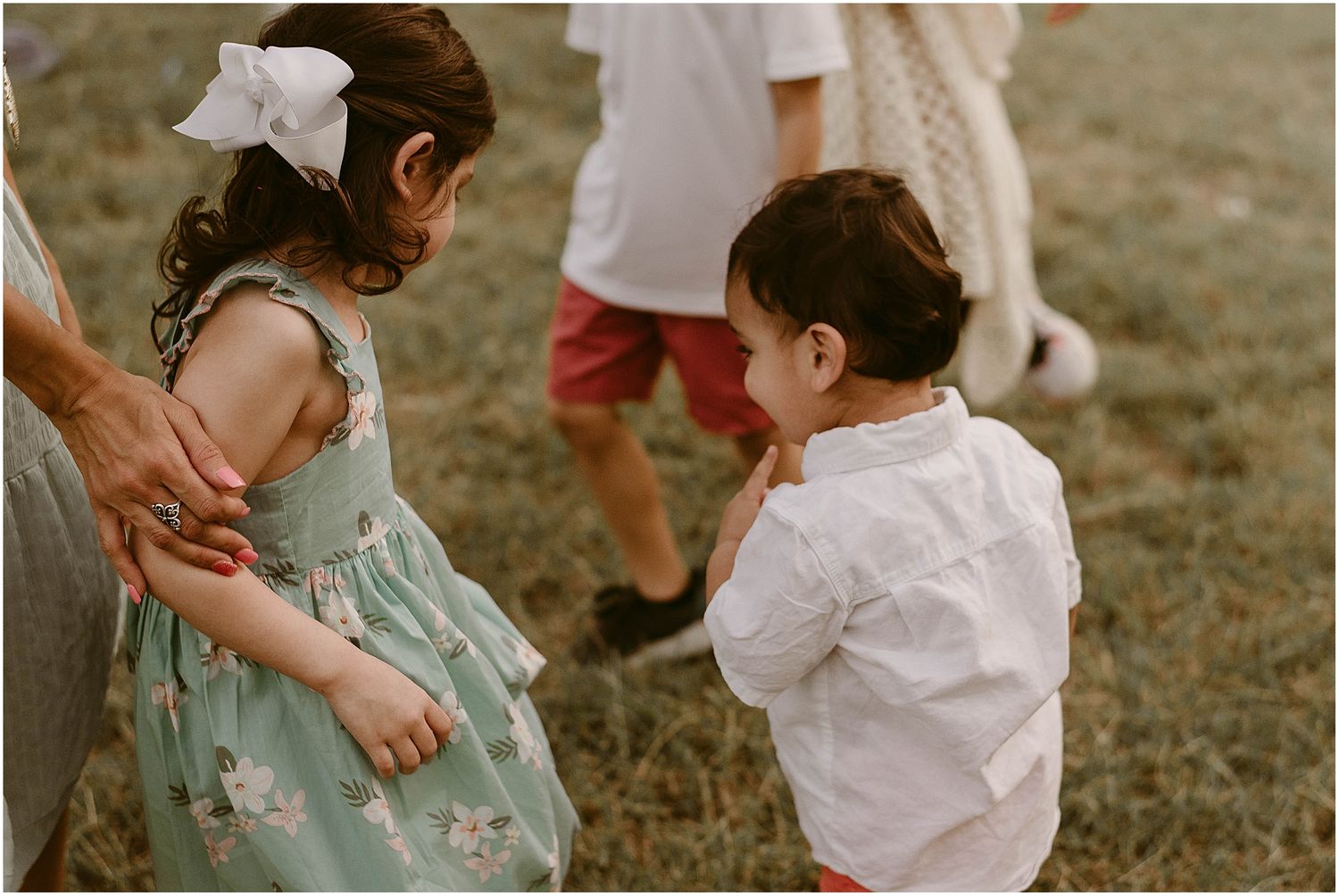 Two small children in summer attire interact while adults stand nearby.