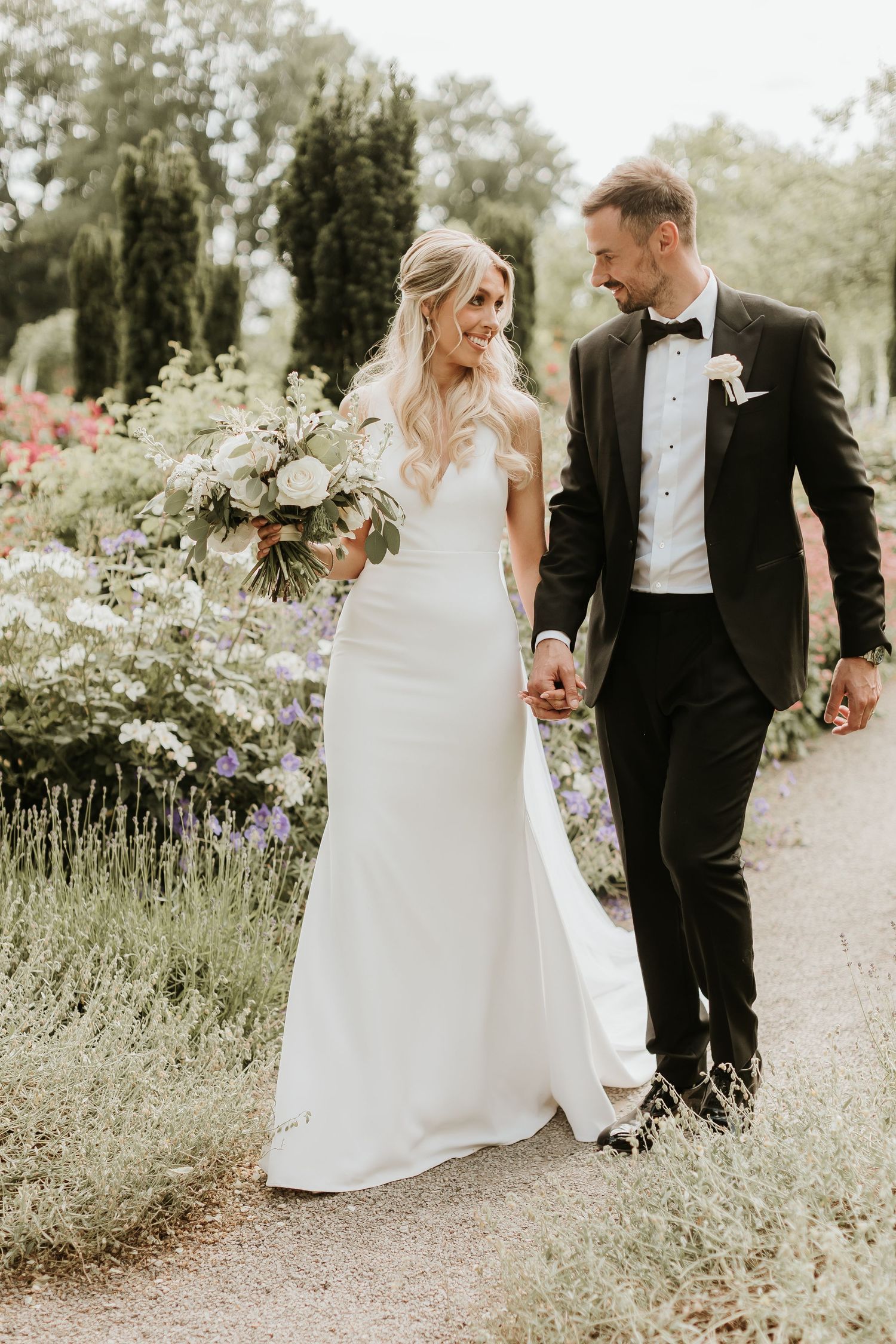 A couple walks hand in hand through a blooming garden pathway in wedding attire.