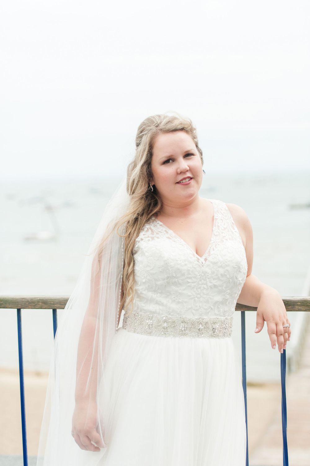 A bride in a flowing white wedding dress poses against a seaside railing.