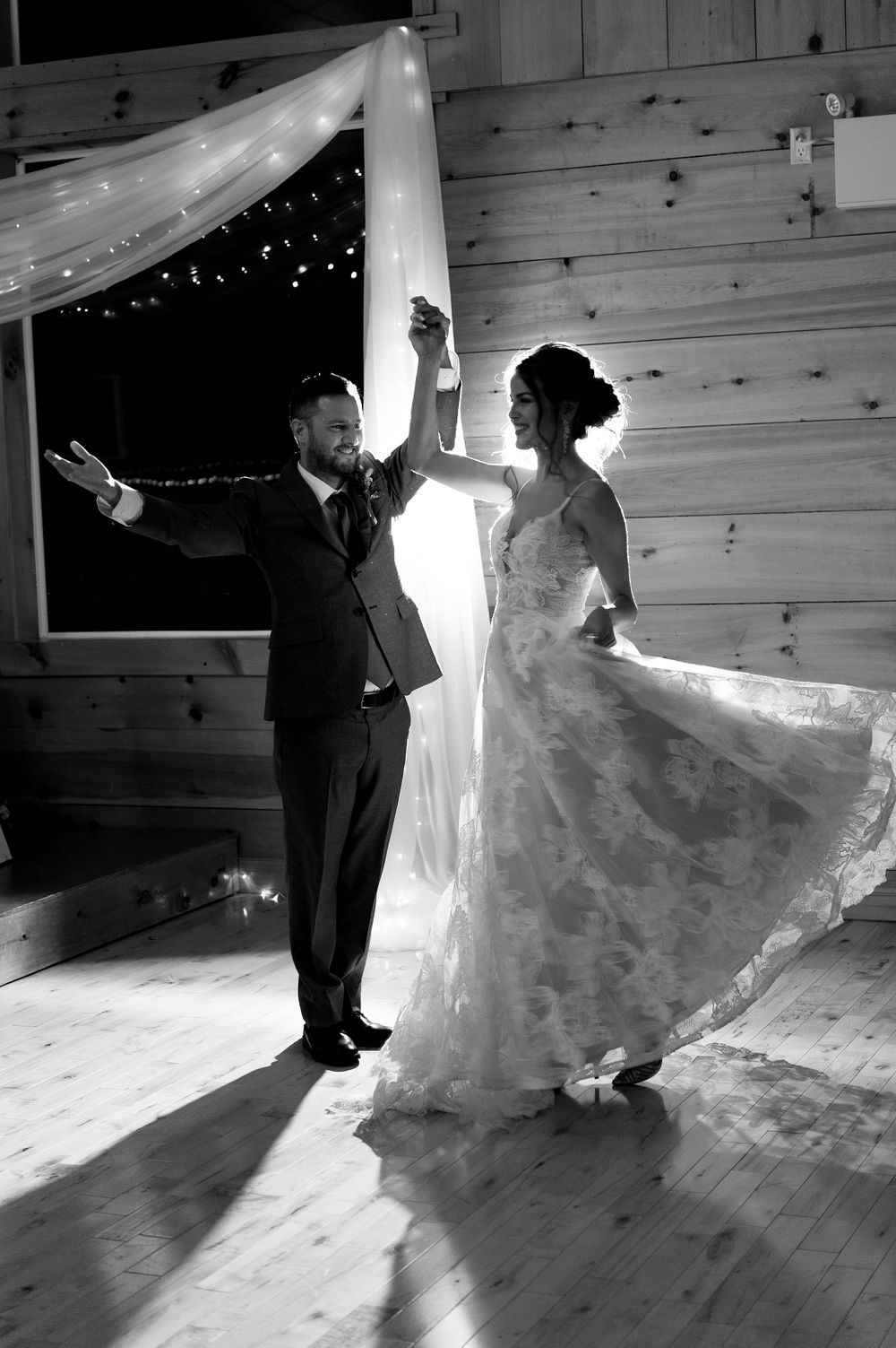 A dramatic black and white wedding dance photo with shadows and string lights in a rustic barn venue.