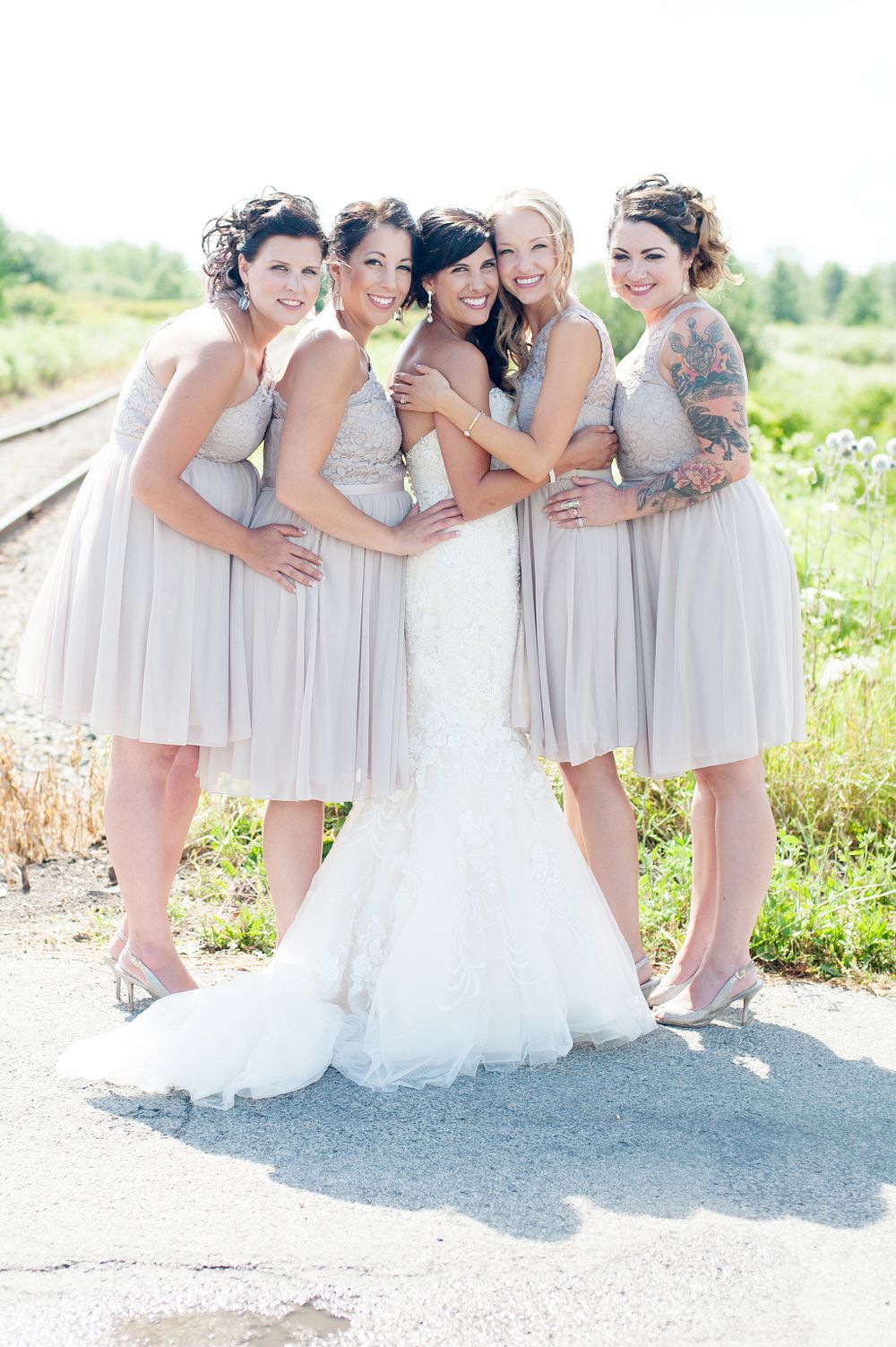 Bridesmaids in light gray dresses gather around bride in white gown outdoors.