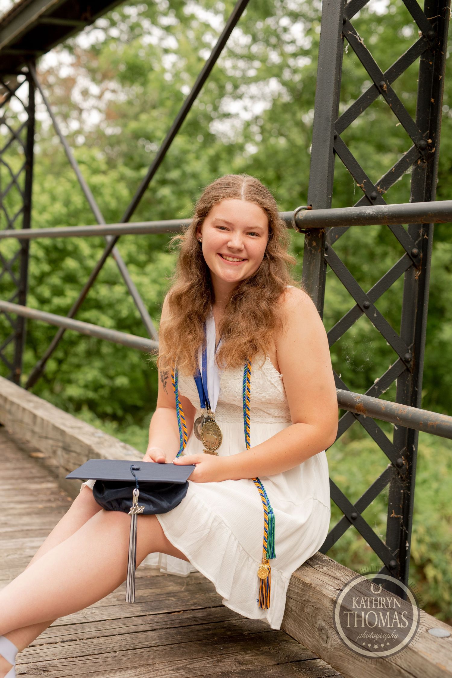 Graduate in white dress and graduation cap sits on wooden bridge during portrait session.