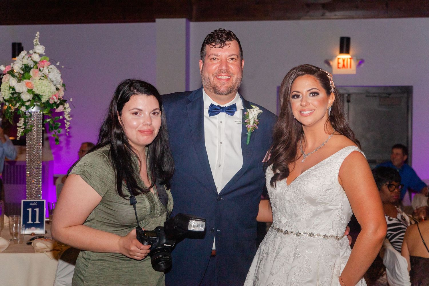 Wedding party members pose together at a reception with floral arrangements and purple lighting.