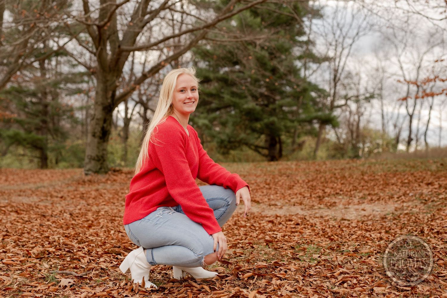 Outdoor autumn portrait crouching in red sweater and jeans among fallen leaves.