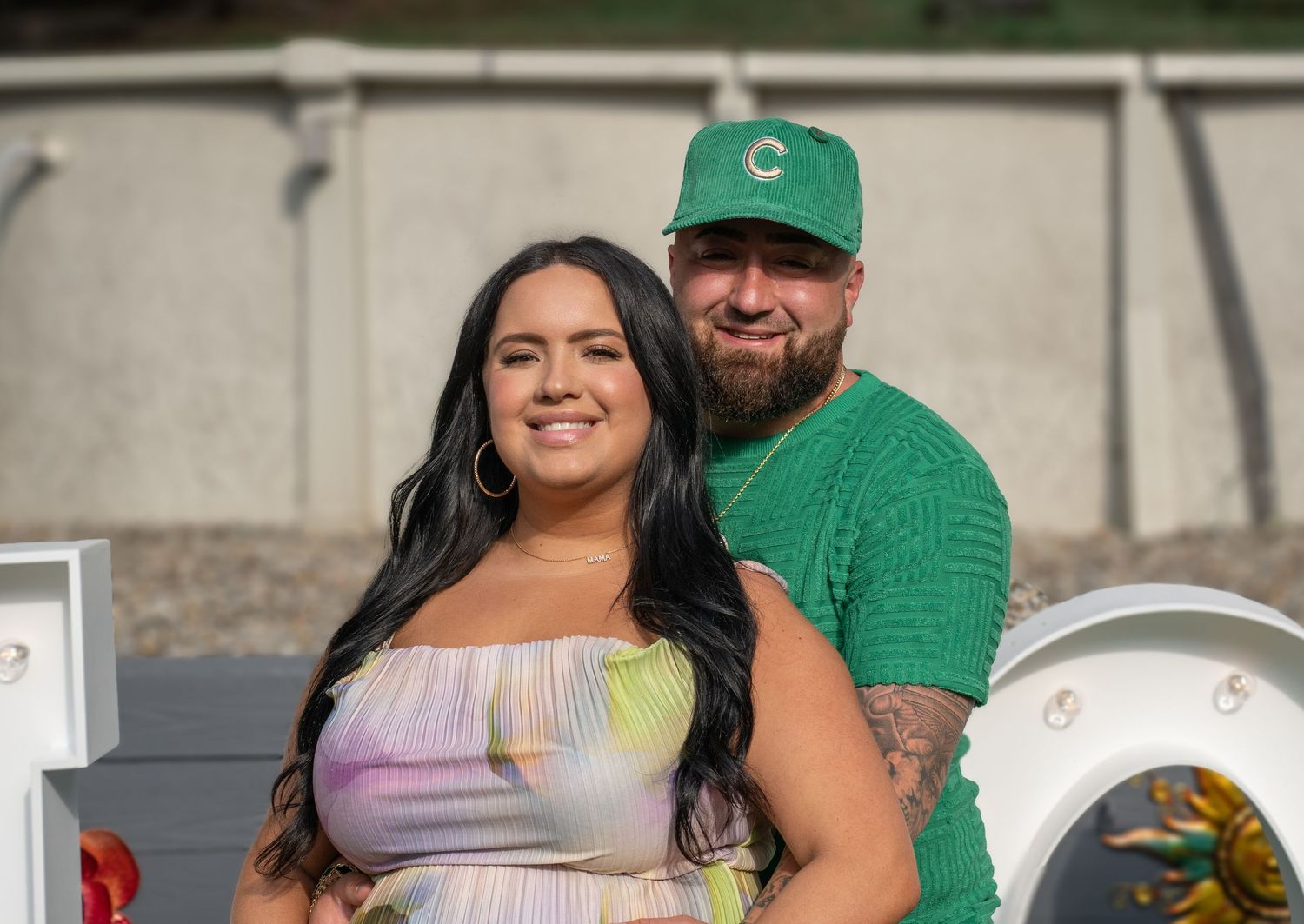 A couple poses together at an outdoor gender reveal party with green and pink decorations.