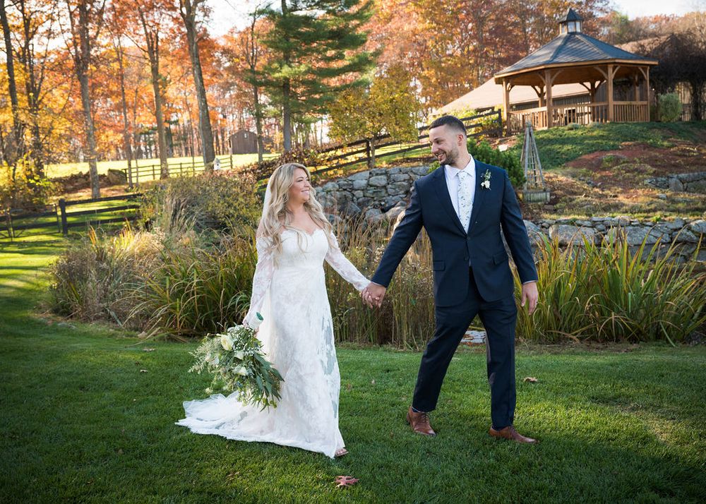 A wedding couple strolls hand in hand through autumn foliage with a rustic gazebo in the background.