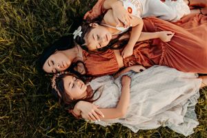 Three people lying in tall grass wearing orange and white dresses at golden hour.