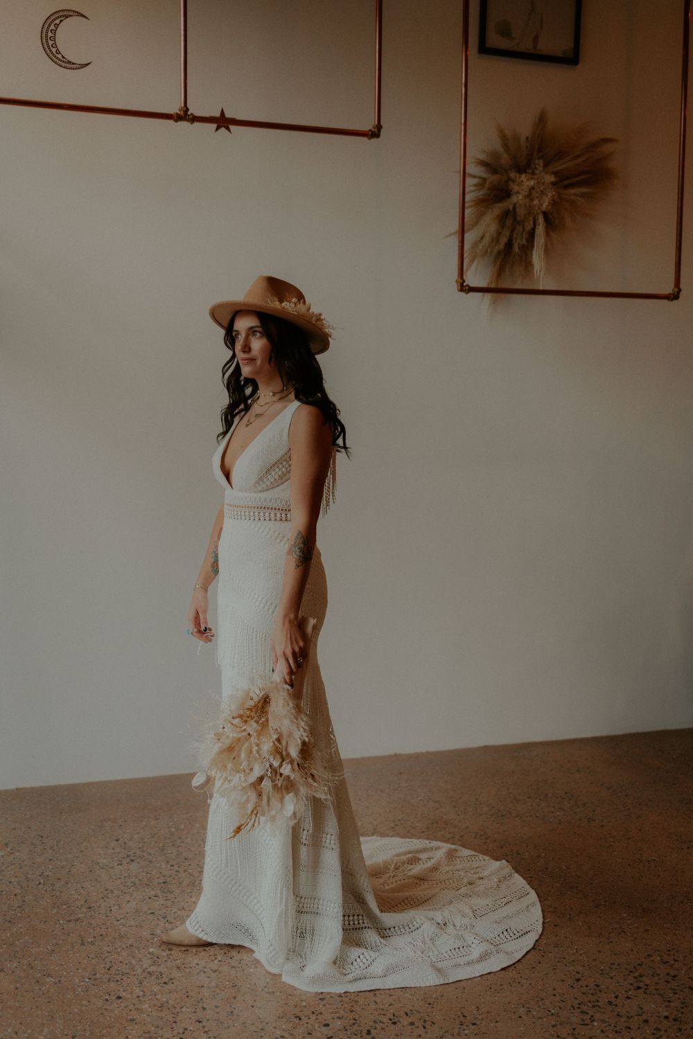 A bohemian wedding dress with a flowing train and brown hat showcased in a minimalist setting with dried pampas decor.