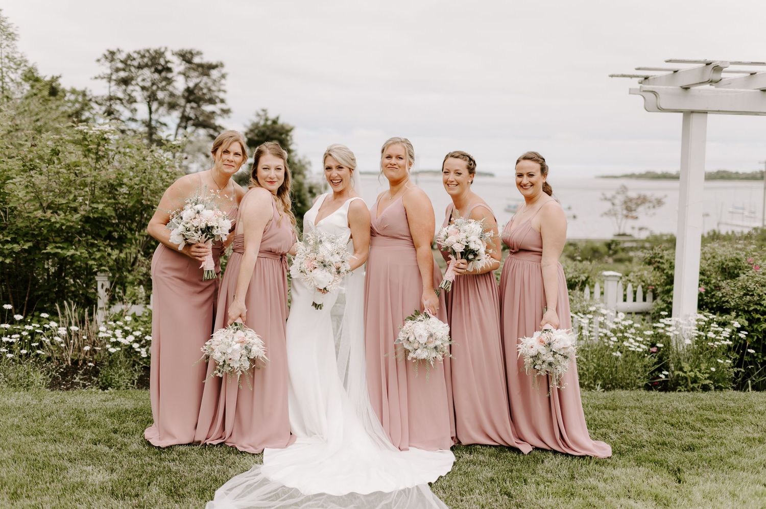 Wedding party poses outdoors with white pergola and coastal backdrop.