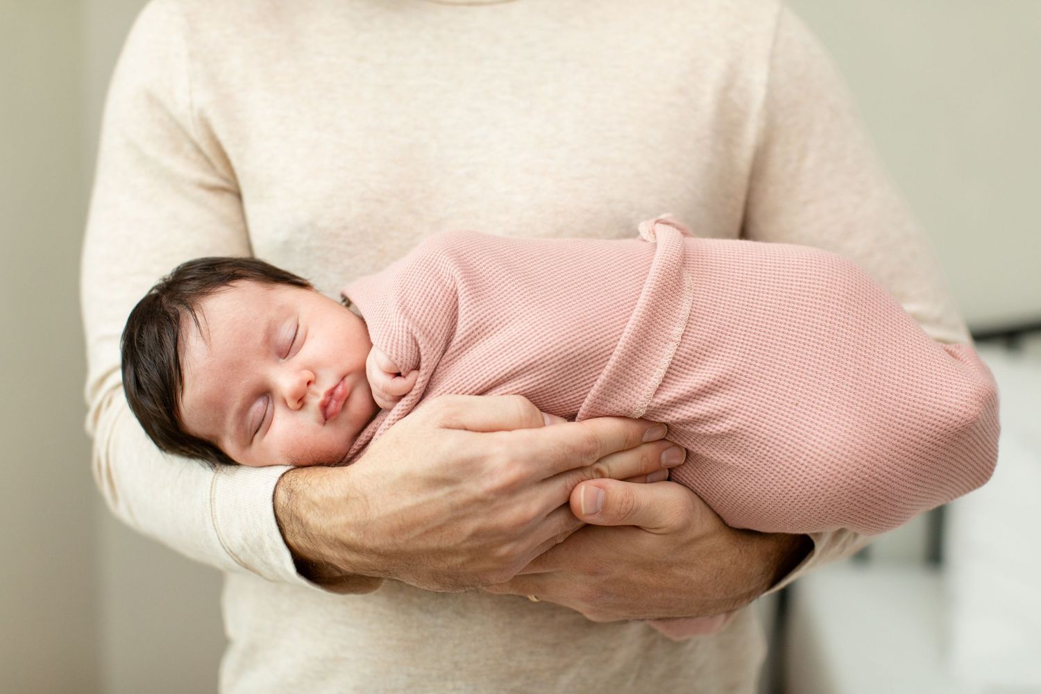 Sleeping newborn baby wrapped in pink knit swaddle is held securely in parent's arms.