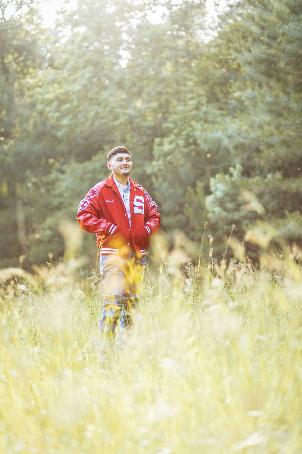 Young man in red varsity jacket standing in sunlit meadow with trees.