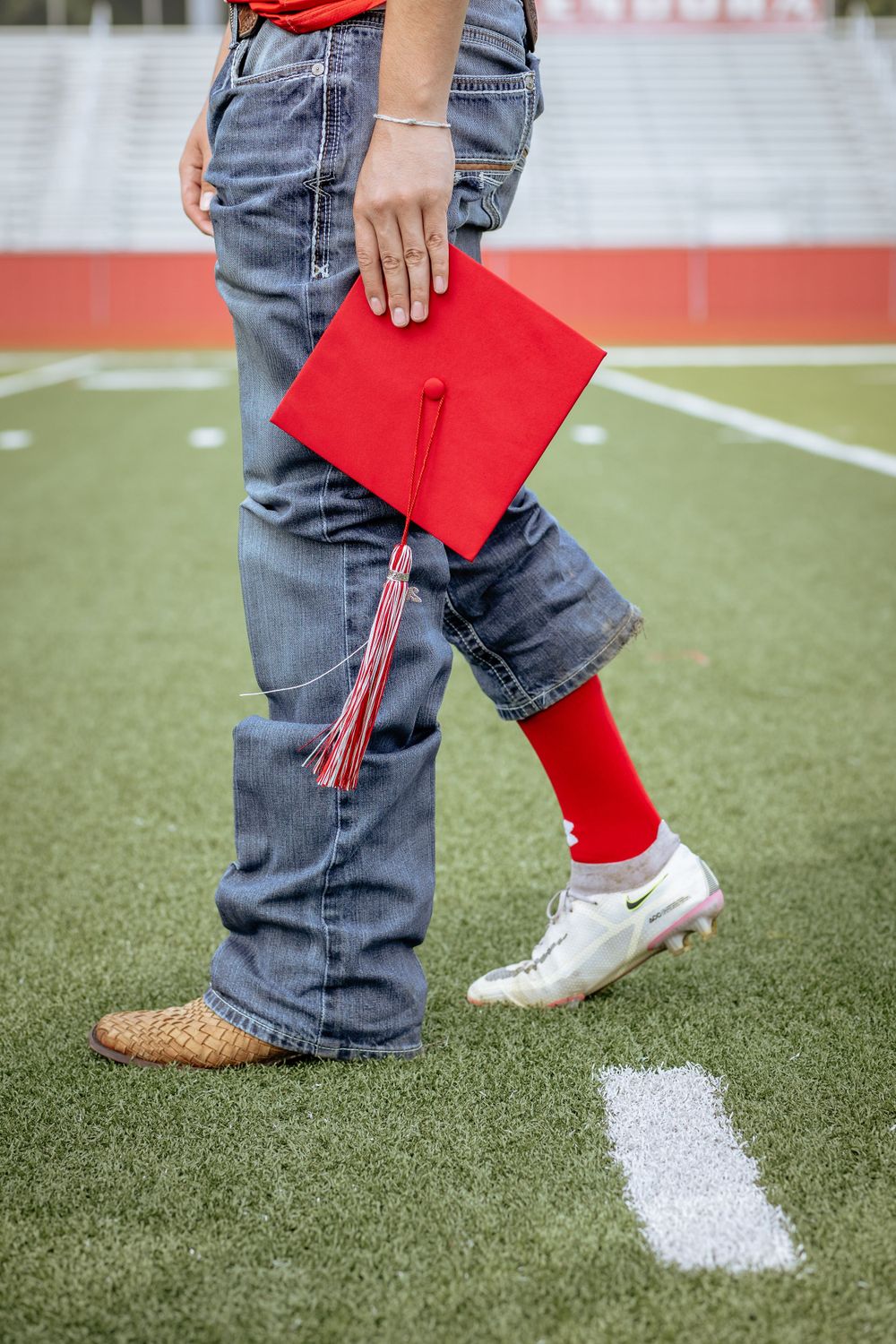 Graduate holding red cap in jeans and soccer cleats on green turf field.
