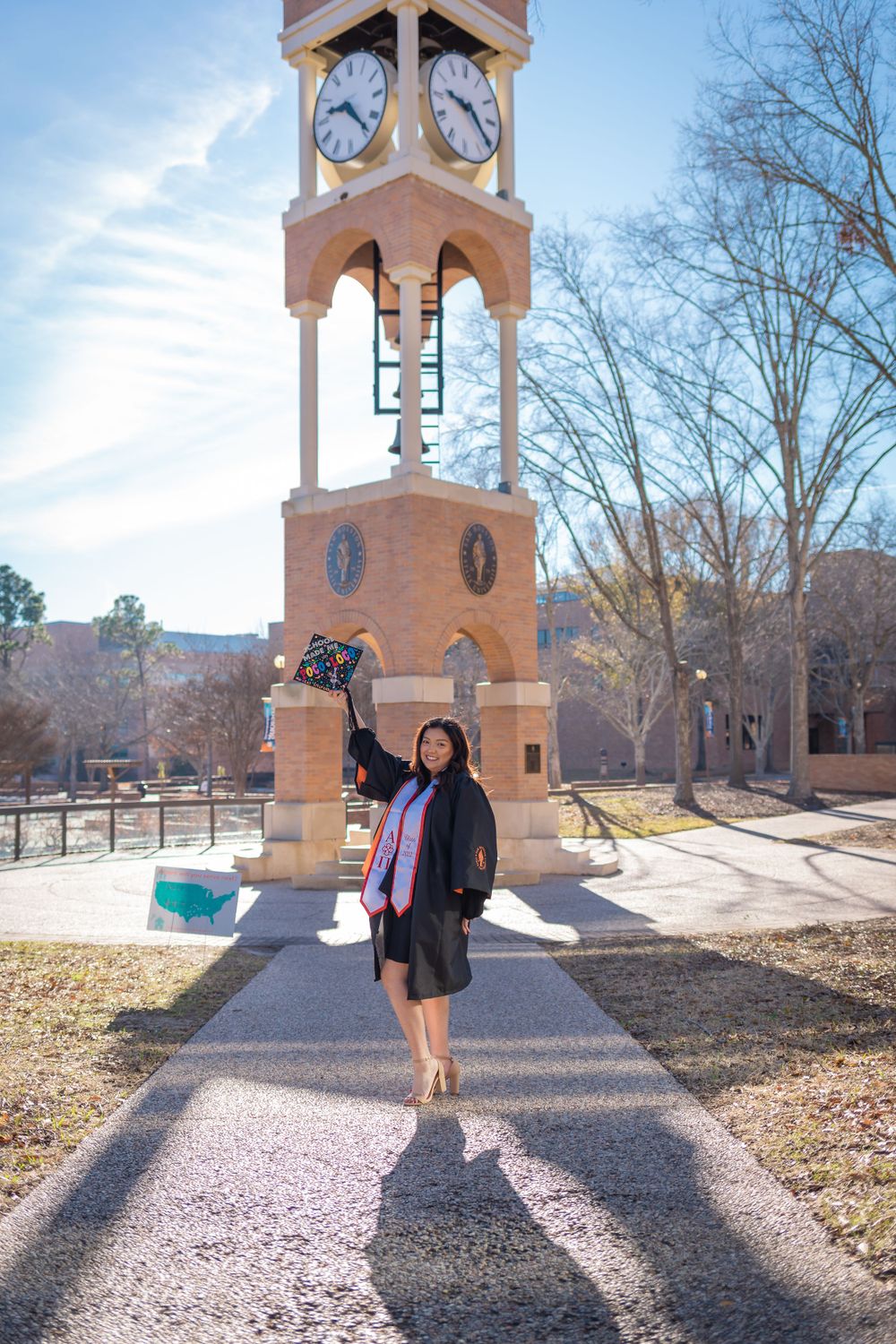 Graduate in cap and gown celebrates by clock tower, tossing decorated cap on sunny campus day.