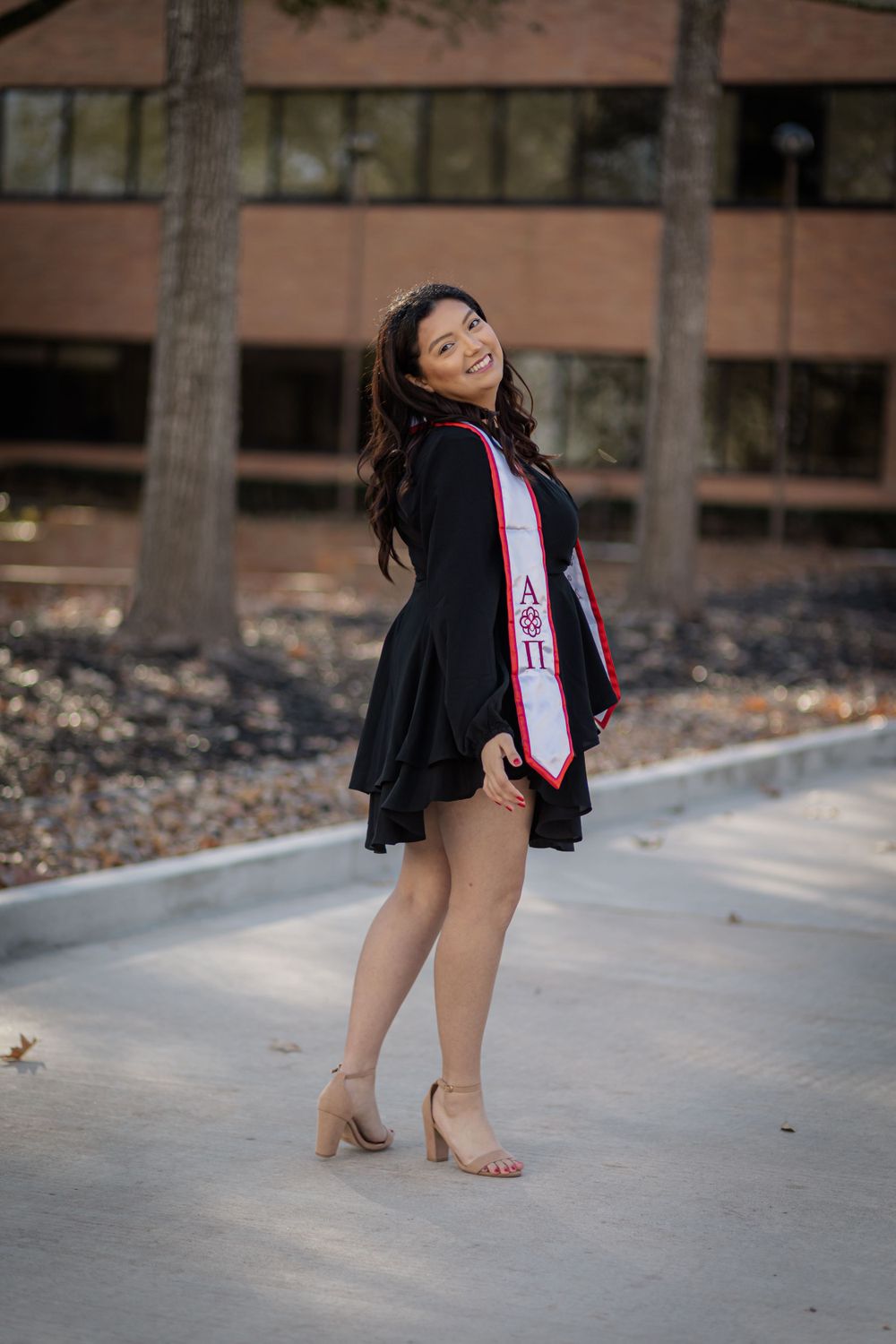 Smiling graduate poses in black gown with Alpha Pi stole on outdoor campus walkway.