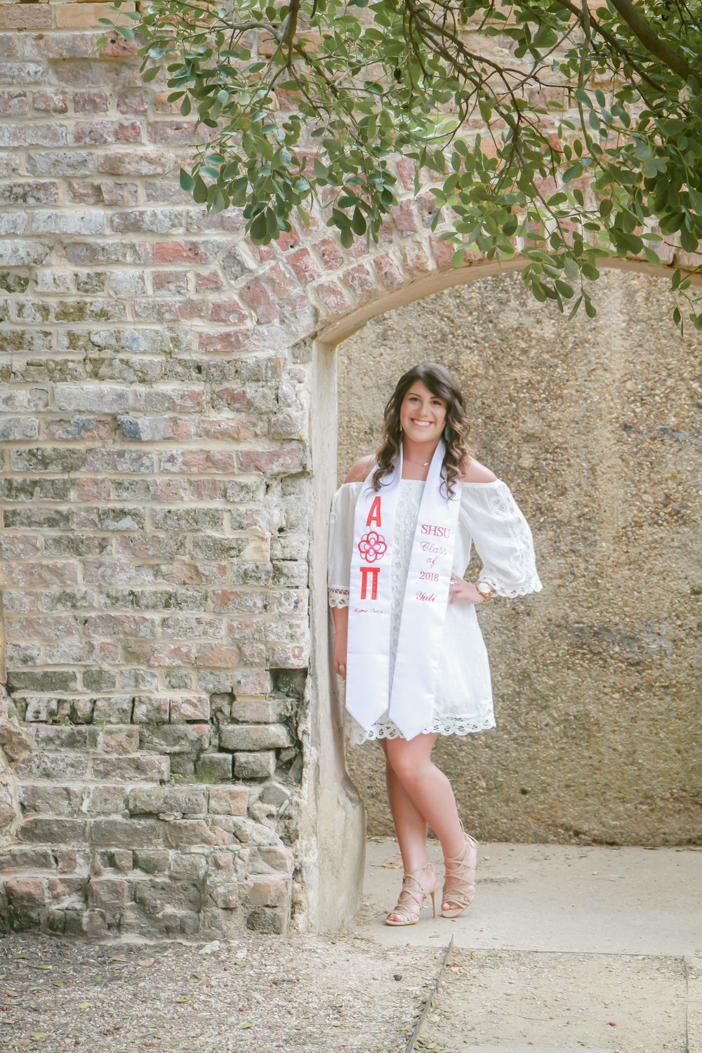 Young woman in white dress with Alpha Pi sorority sash poses under stone archway for graduation photos.
