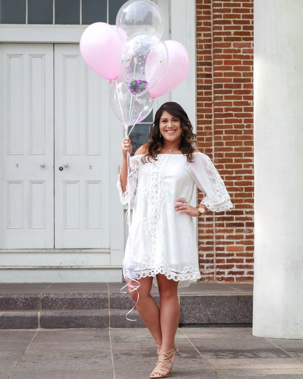 Young woman in white lace dress holding pink and clear balloons outside brick building.
