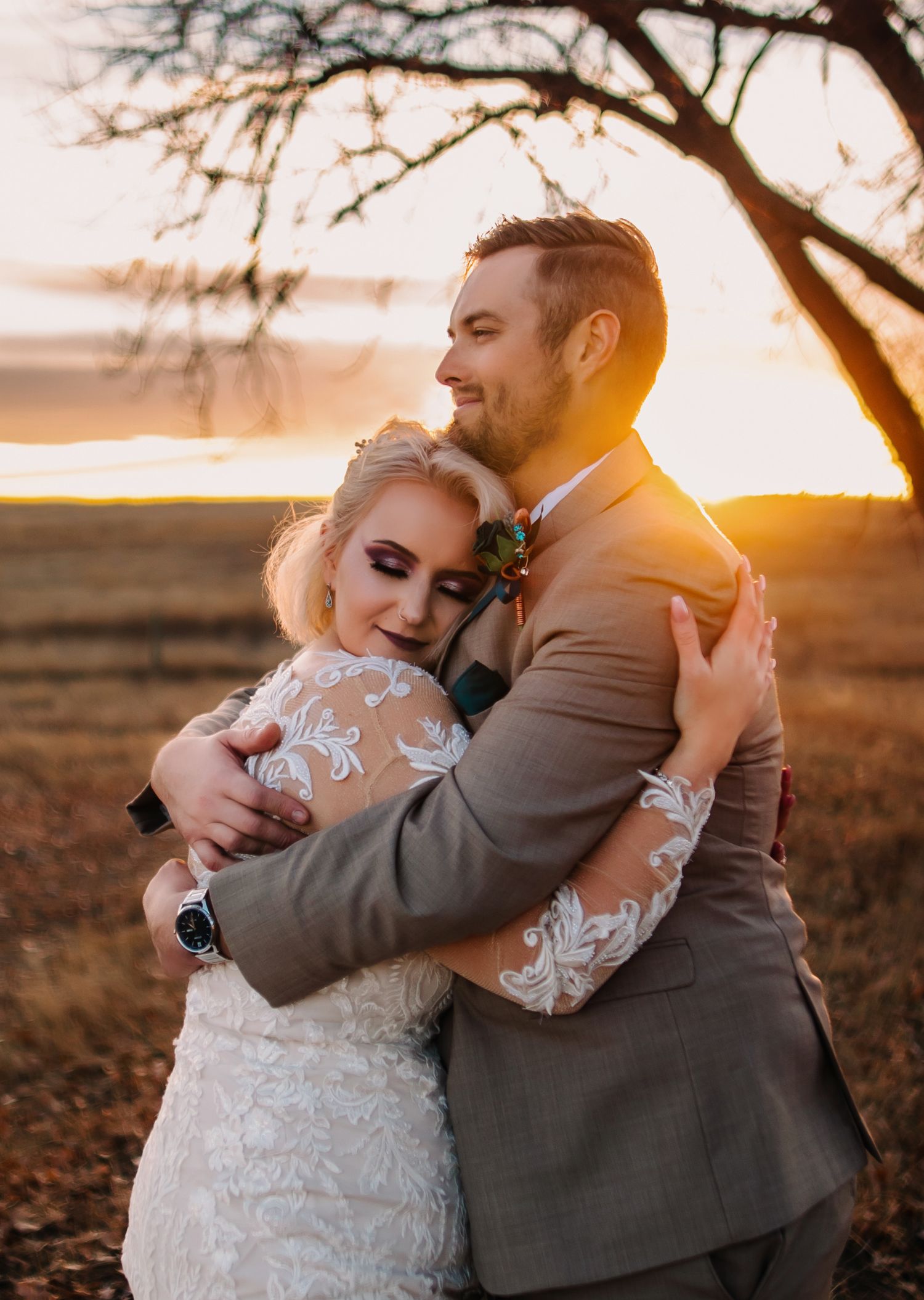 A couple embraces during golden hour sunset under a tree branch, with the bride wearing a lace sleeved wedding gown.