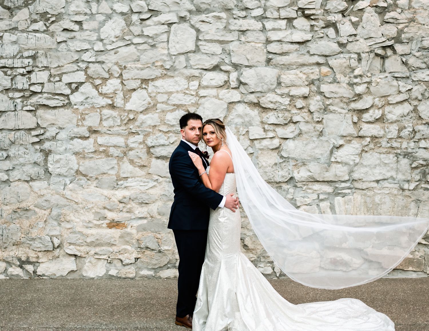 A flowing wedding dress train creates elegant movement against a stone wall backdrop.