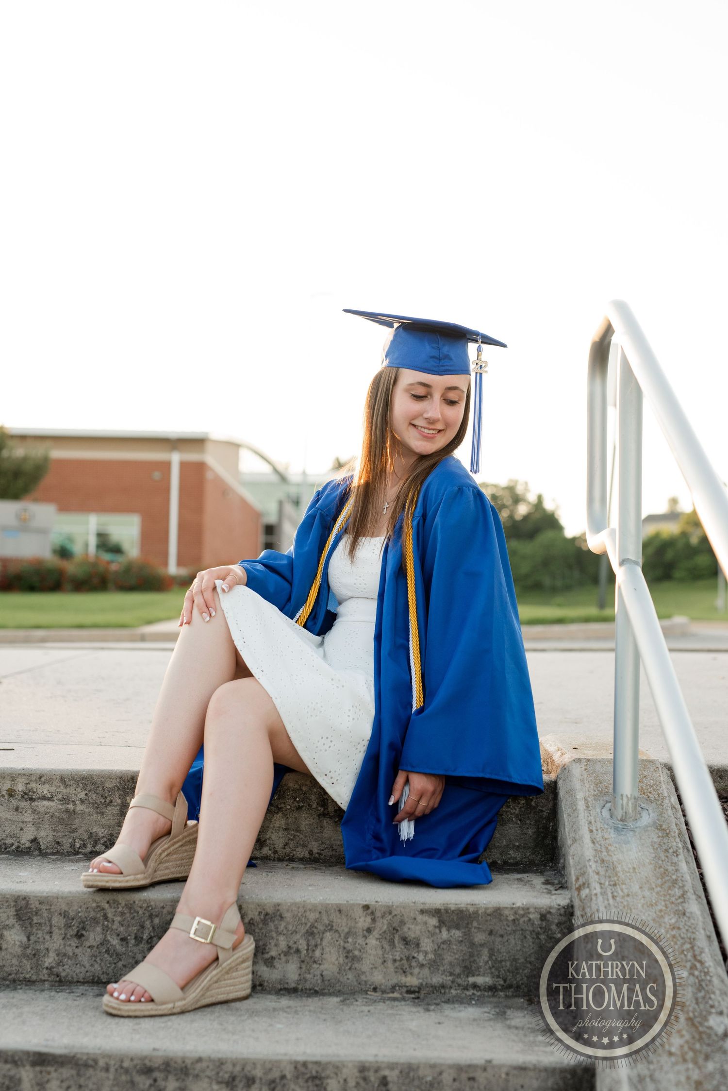 Graduate in blue cap and gown sits casually on concrete steps during photo session.