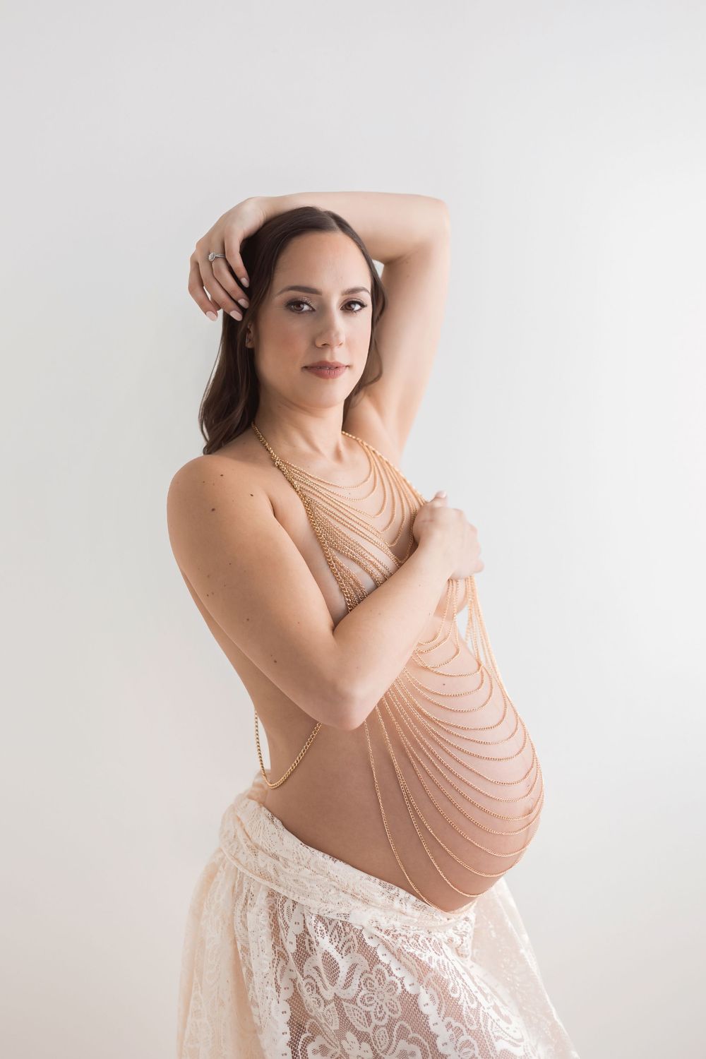 Pregnant woman poses elegantly in body chains and lace skirt against a white studio background.