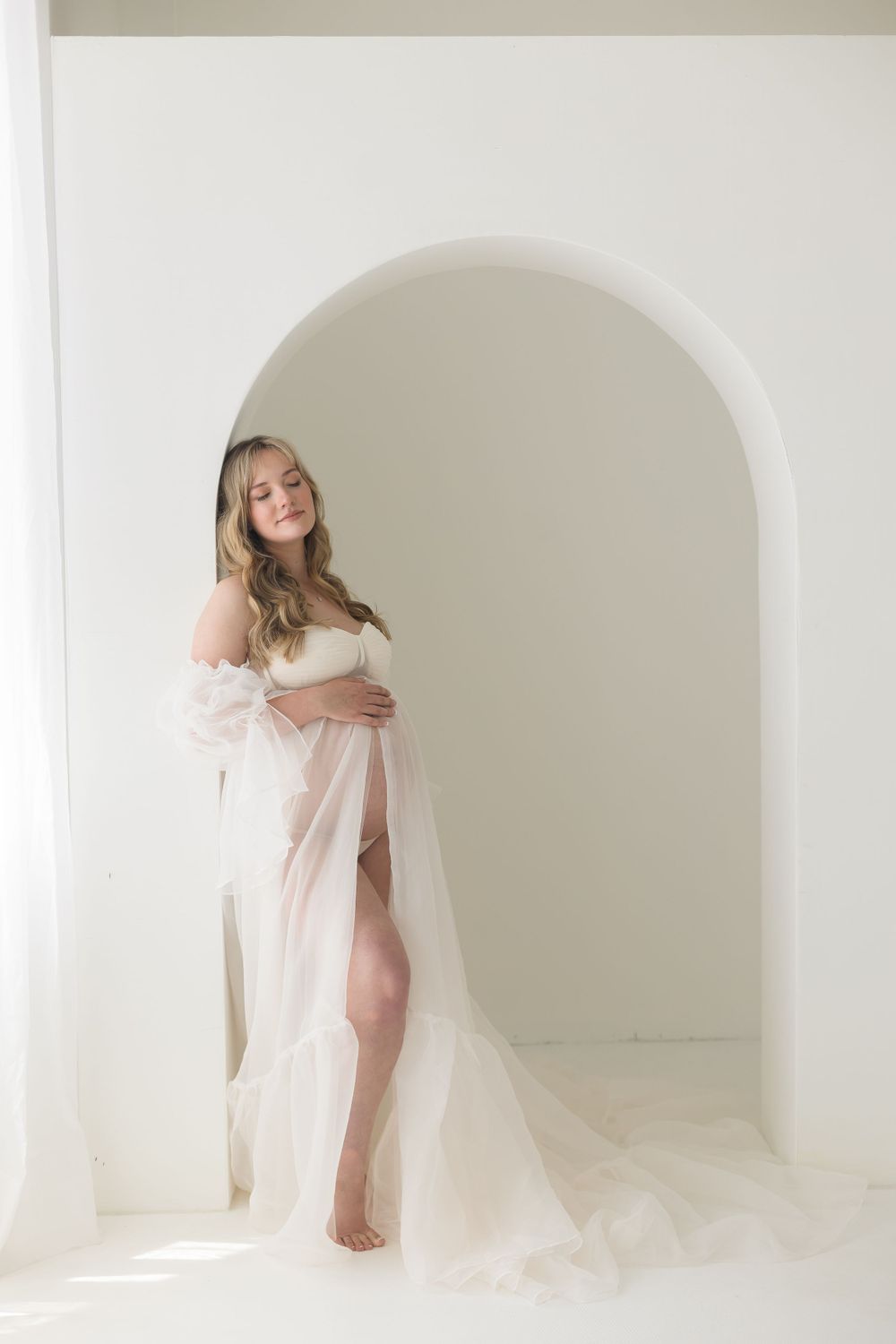Pregnant woman in sheer white tulle off-shoulder maternity gown posing in white arched doorway.