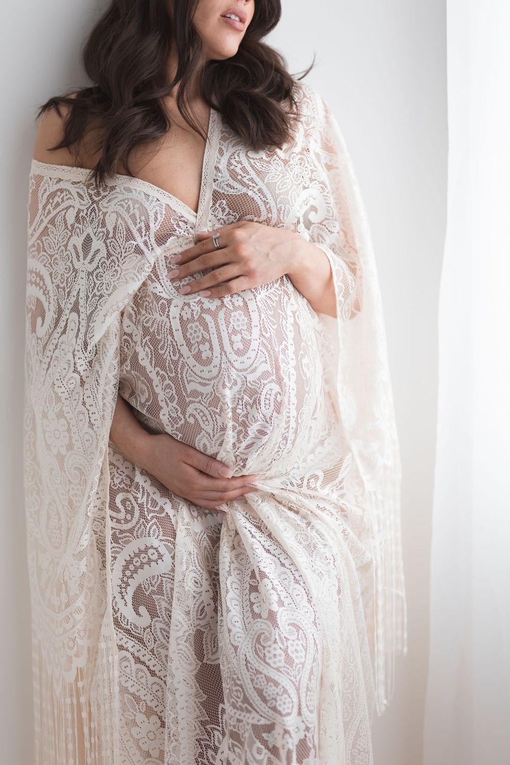 Pregnant woman wearing sheer white lace cape maternity dress cradling baby bump in bright studio.