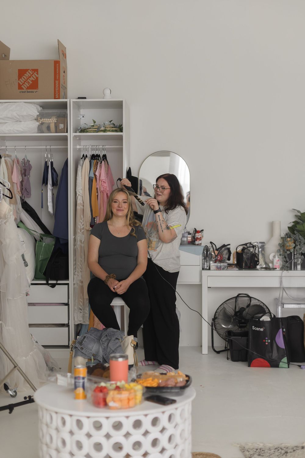 Hair stylist blow-drying seated womans hair in bright photography studio with wardrobe rack.