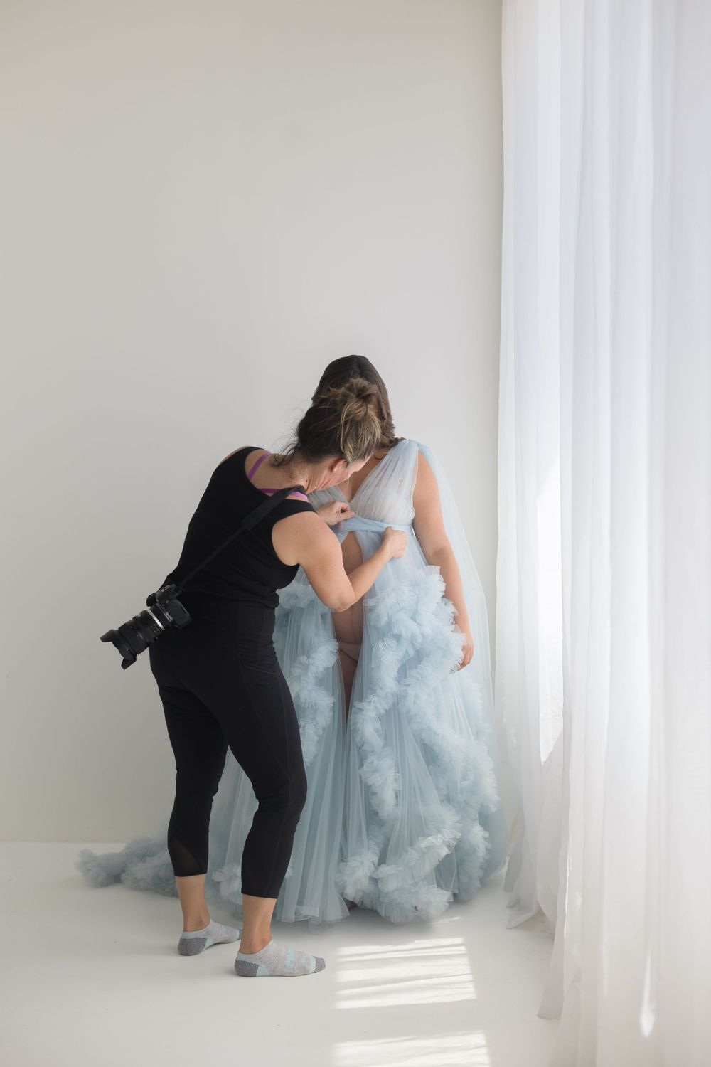 Photographer adjusting light blue cloud tulle maternity gown on pregnant woman near studio window.