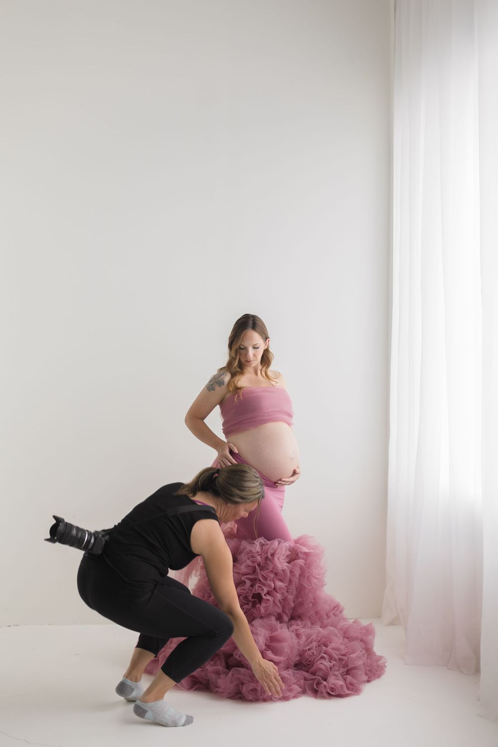 Photographer crouching to arrange pink tulle maternity skirt on pregnant woman in white studio.
