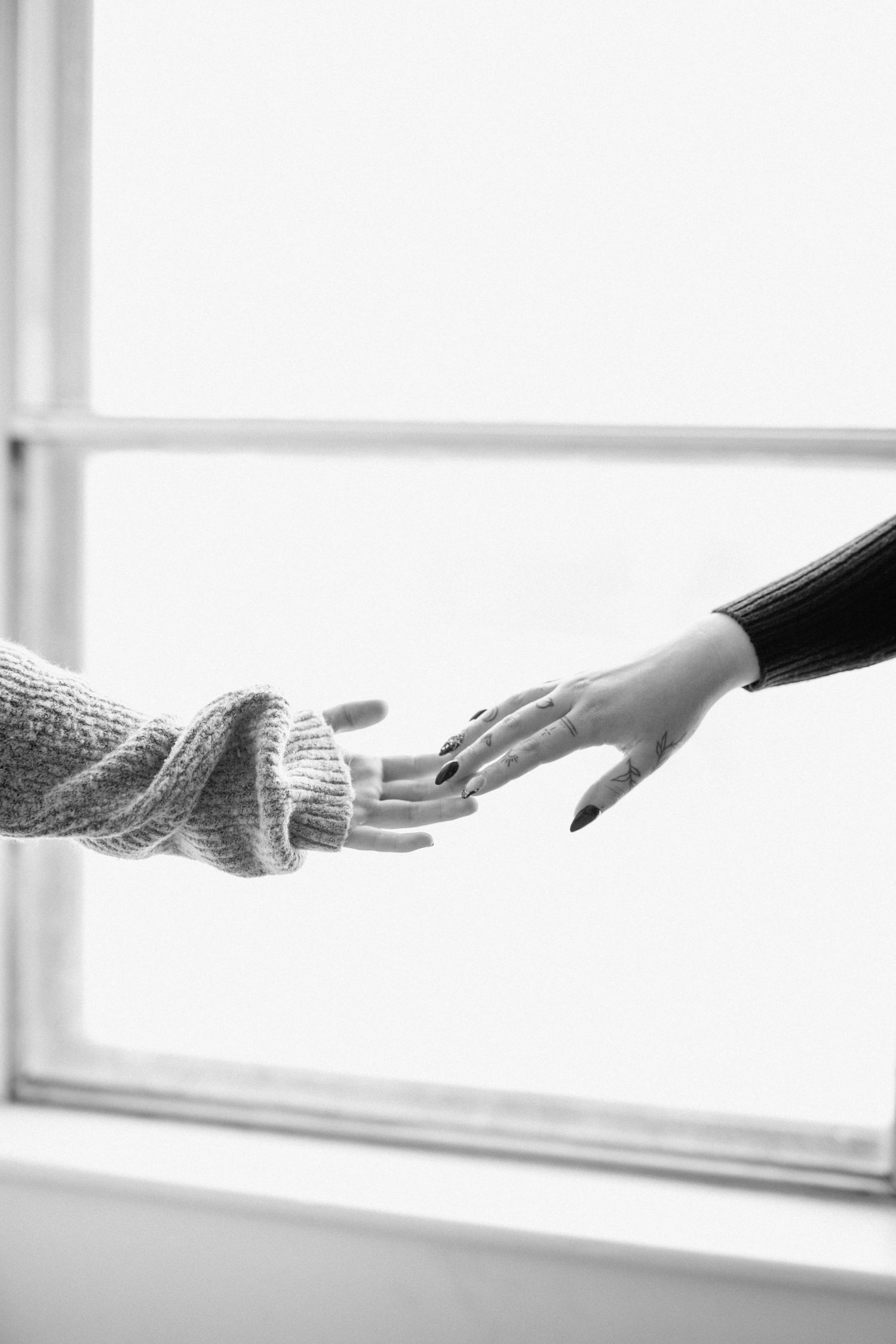 Black and white image of two hands reaching toward each other near a window.