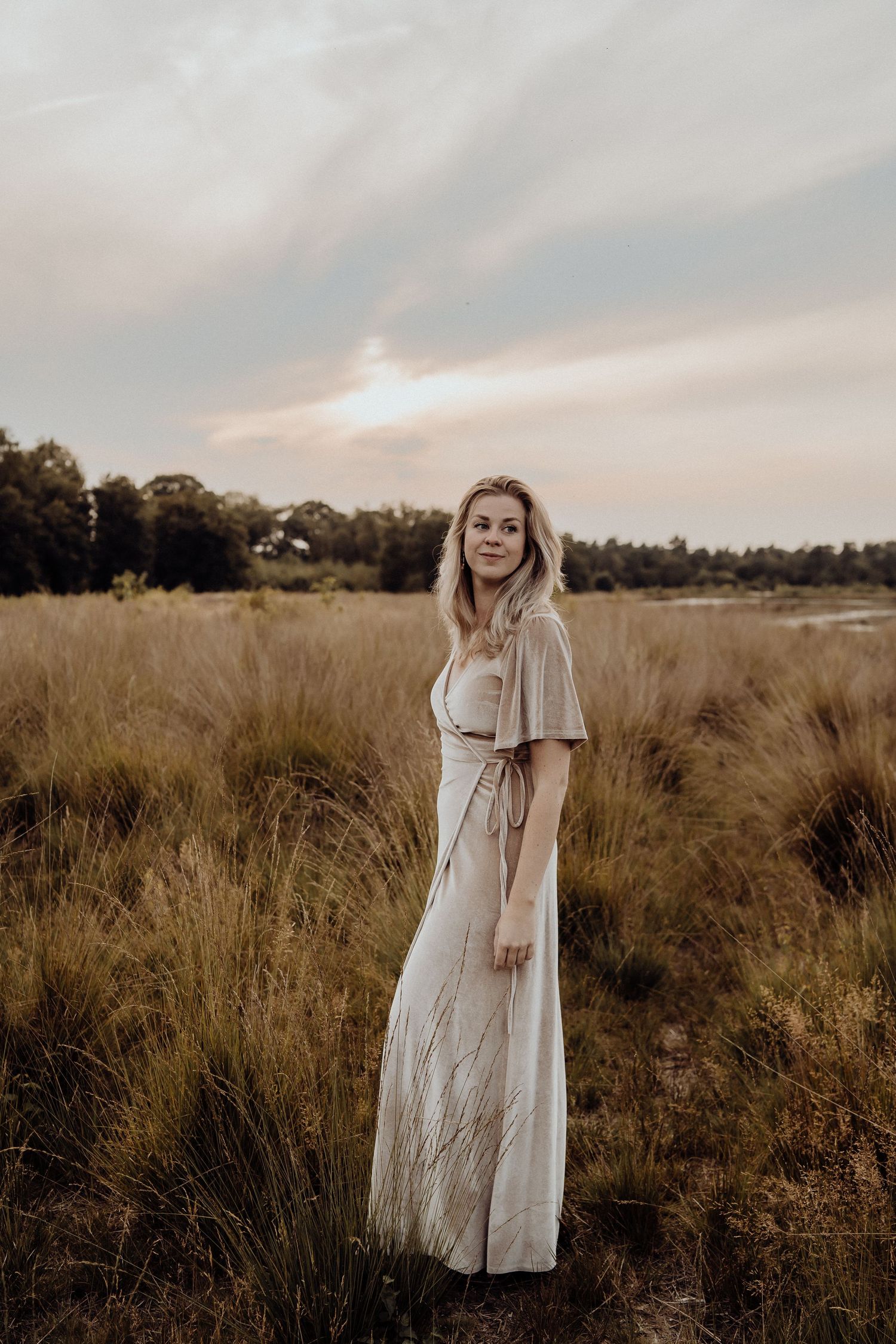 Woman in flowing white dress stands alone in tall grass at sunset.