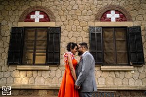 A couple in formal attire stands intimately in front of a stone building with cross-adorned windows.