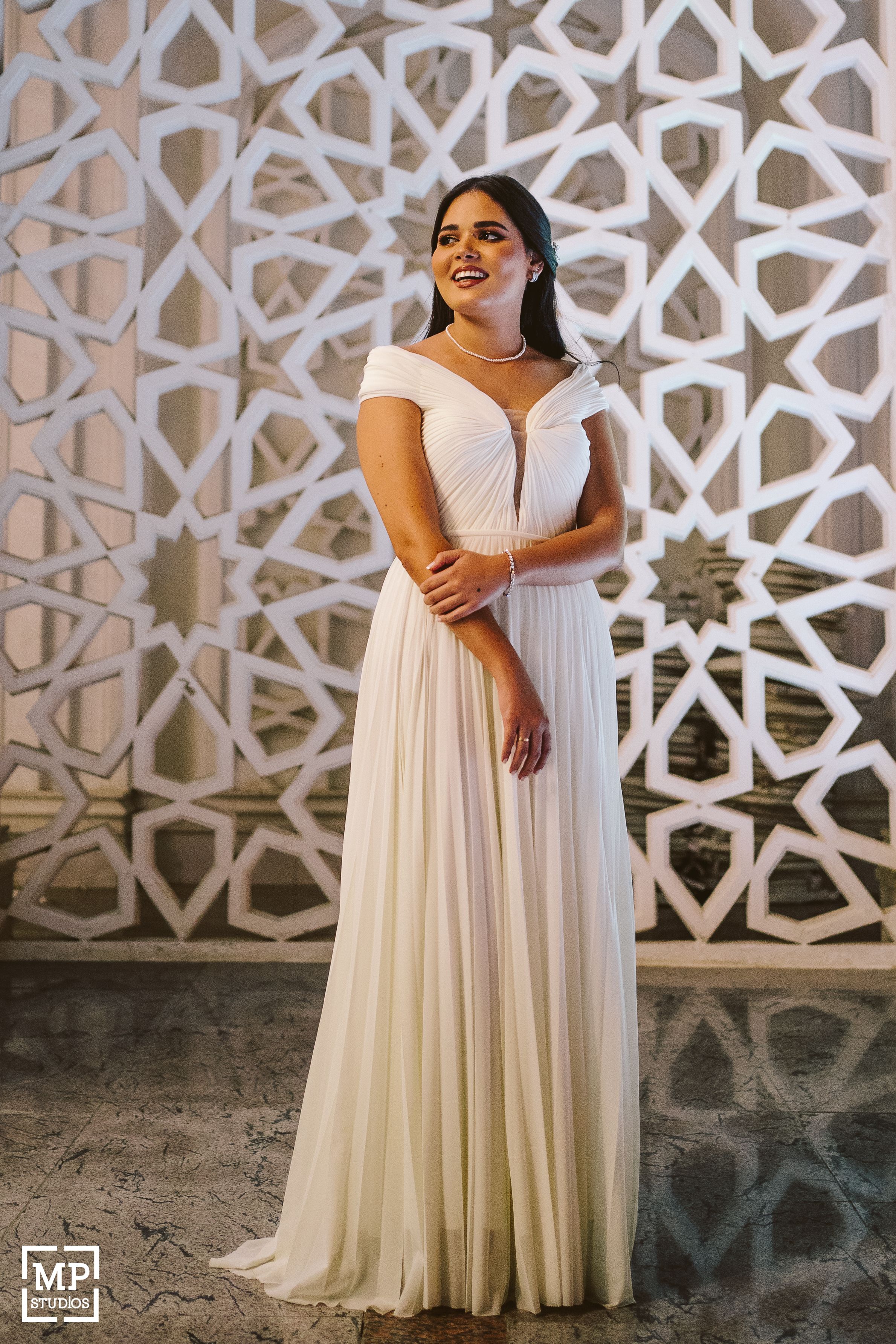 A bride in an elegant off-shoulder white gown poses against a geometric patterned wall.