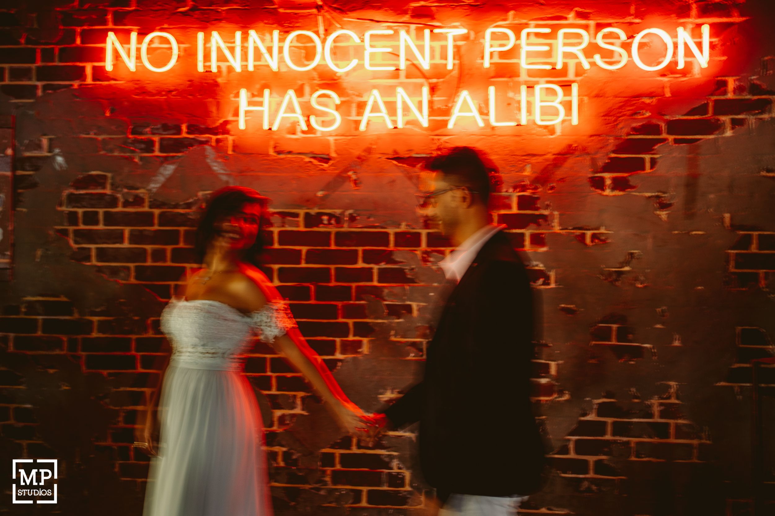 A couple poses beneath a red neon sign against a brick wall.