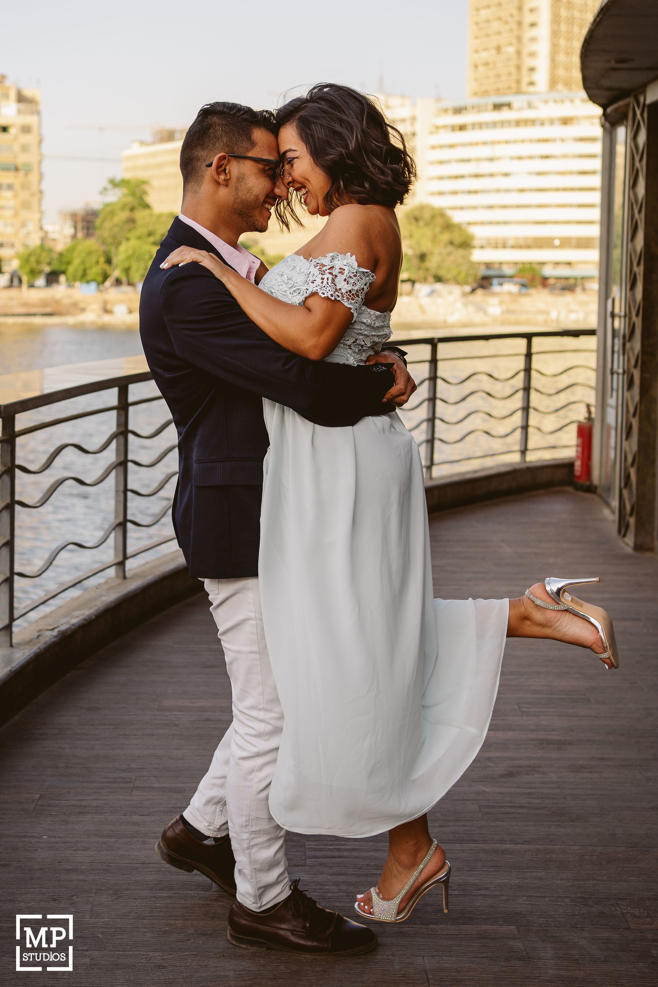 A romantic couple embraces on a waterfront deck, with the bride wearing a flowing white dress and the groom in a dark blazer.