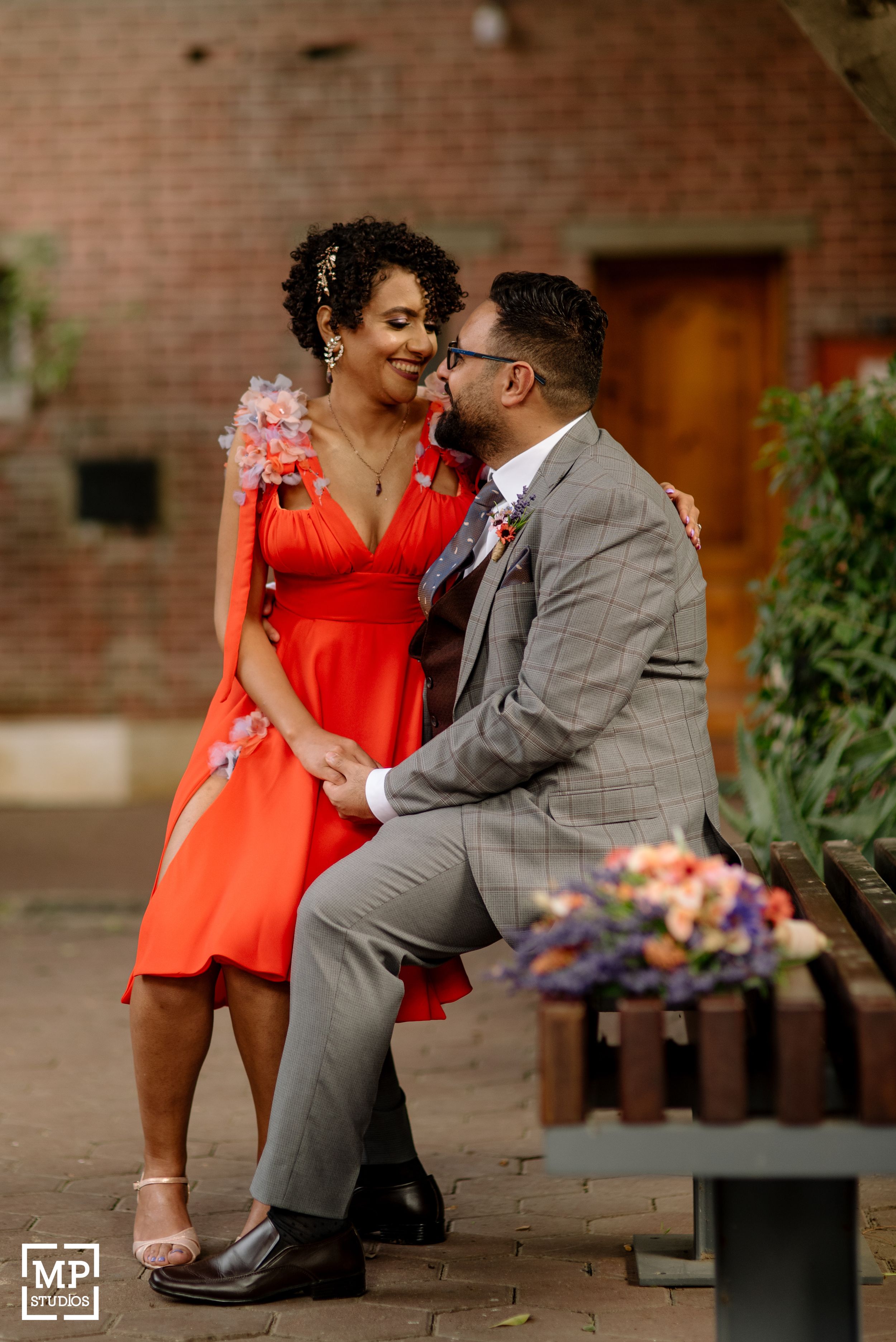 A couple shares a tender moment on a bench, with the woman in a vibrant red dress.