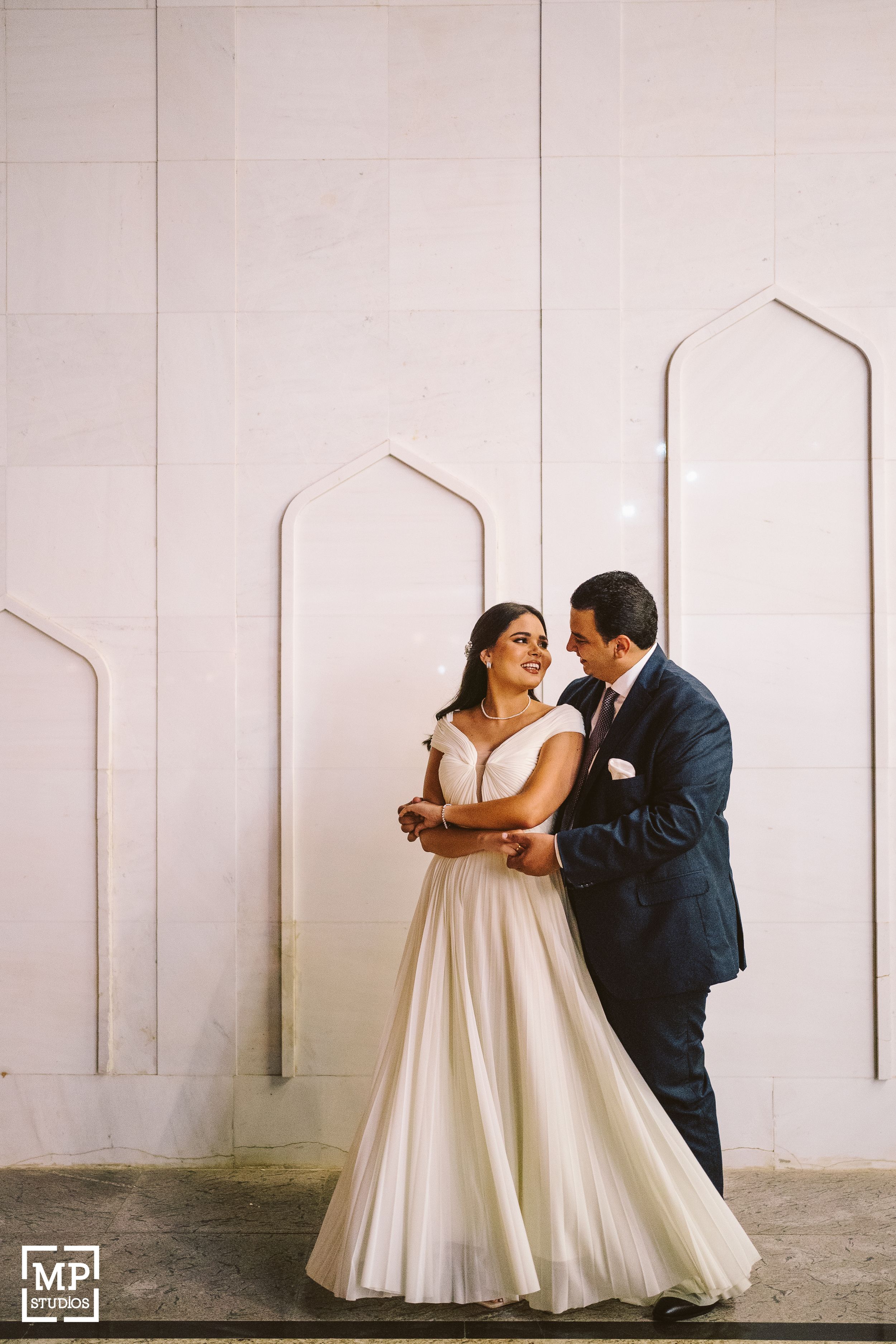 A romantic wedding portrait against a white wall with architectural arches.