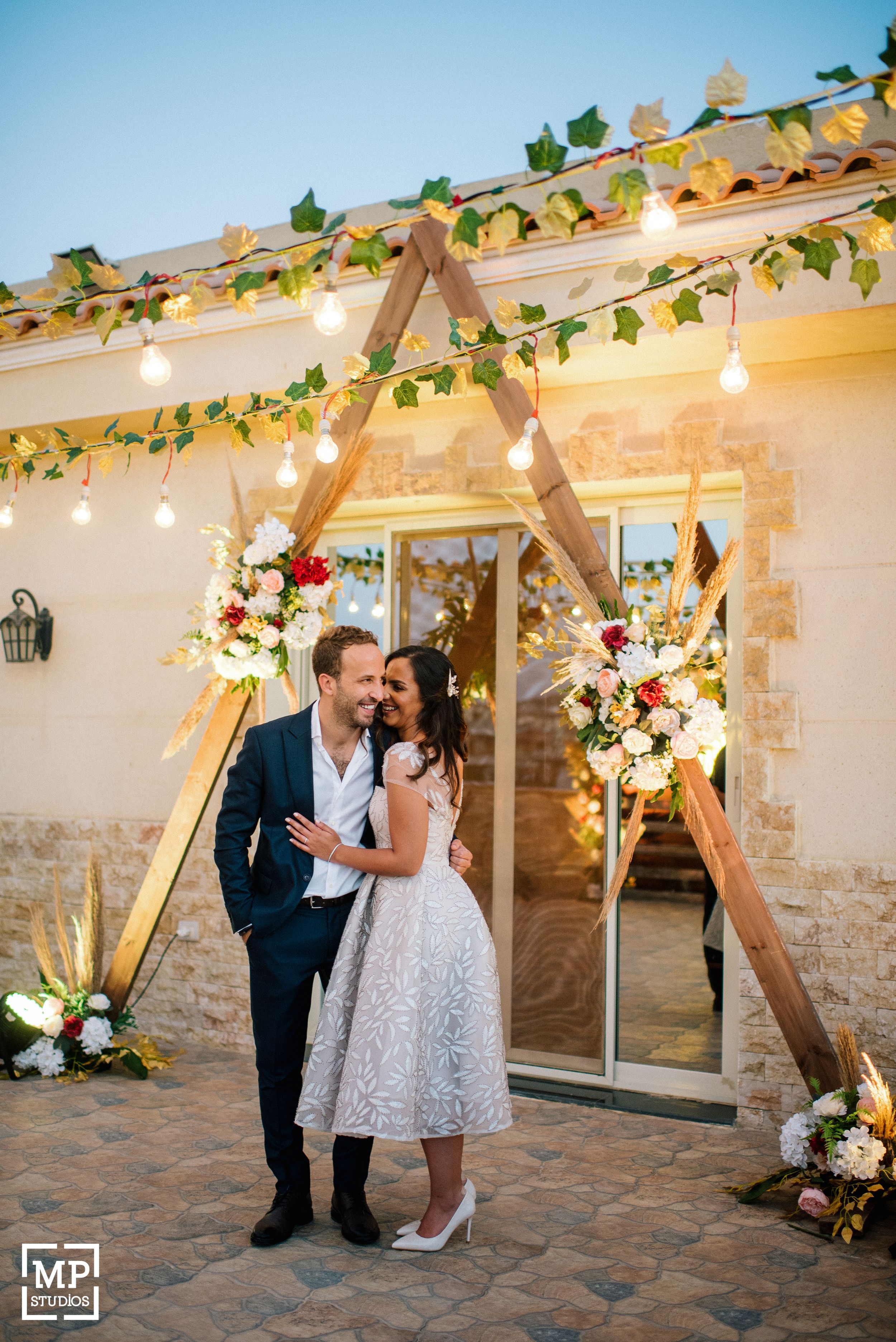 Wedding couple under wooden arch with string lights and floral decorations against stone wall.