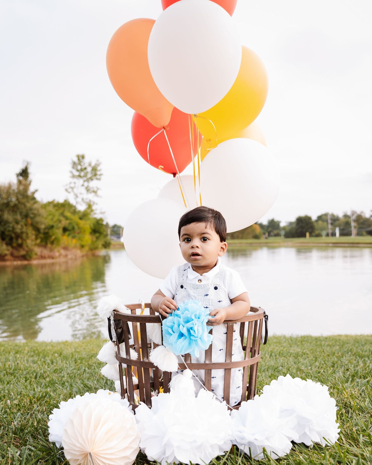 Adorable first birthday photoshoot by a lake with colorful balloons and a rustic wooden basket decoration.
