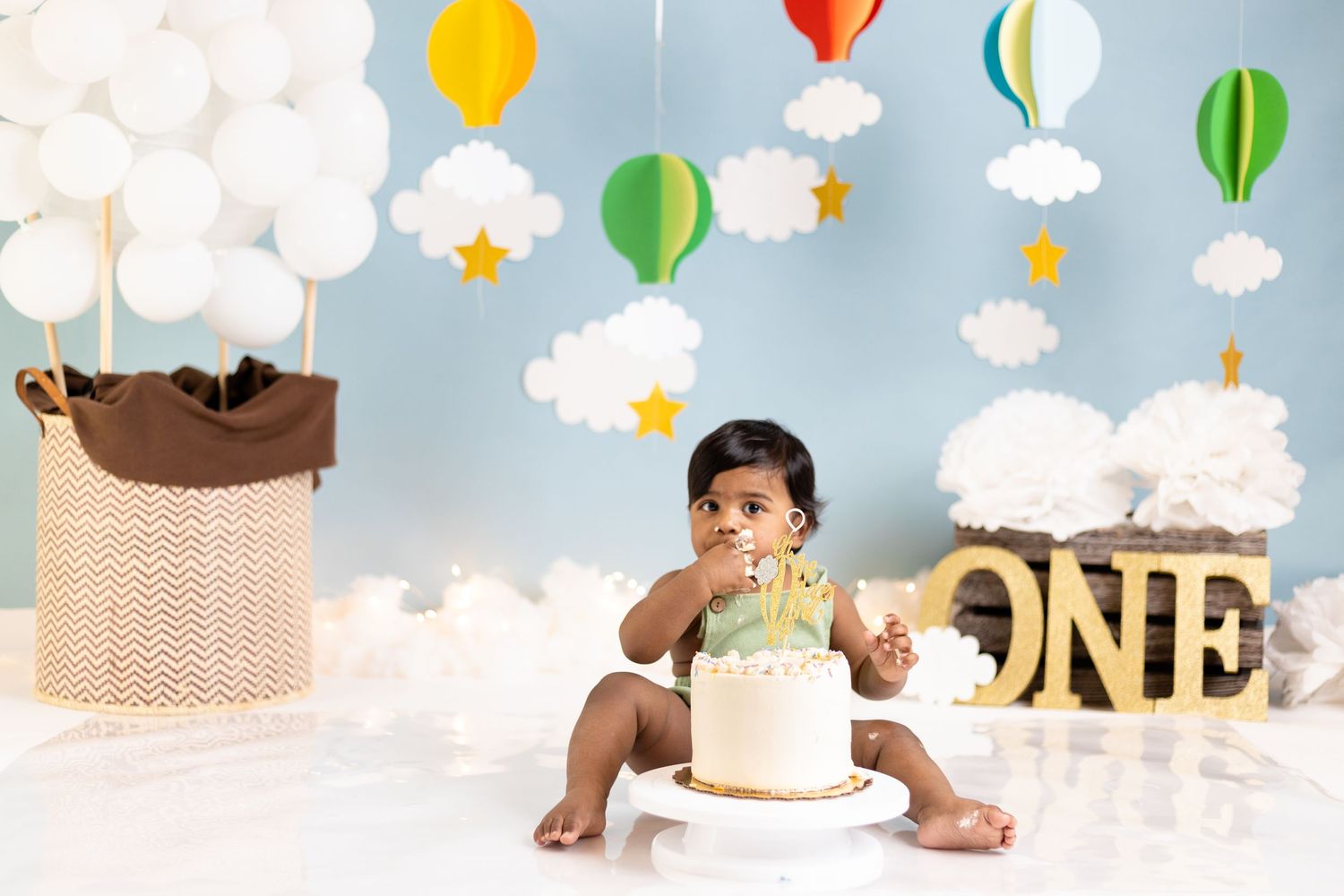 First birthday party setup with colorful hot air balloon decorations, clouds, and a cake against blue background.