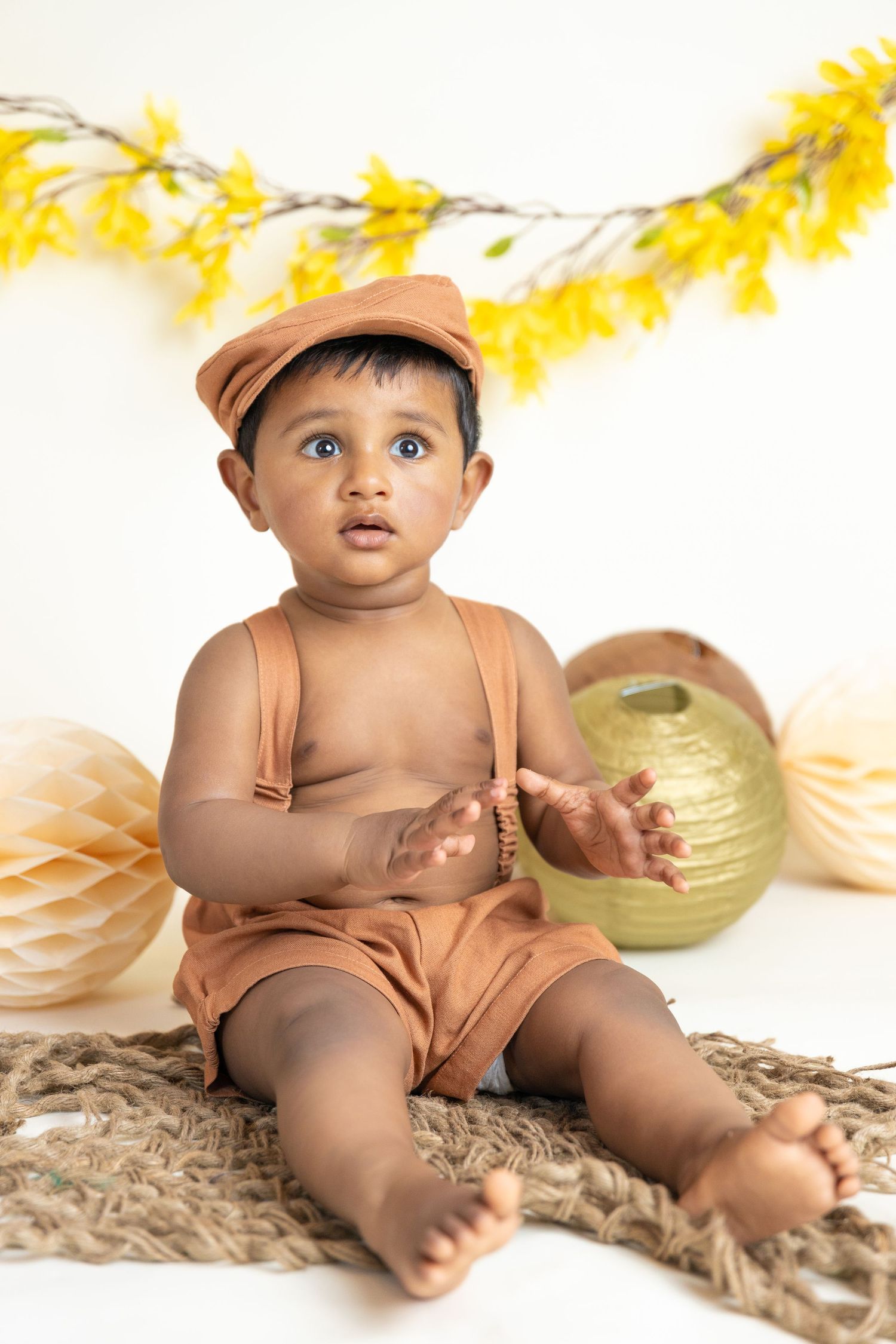 Baby in tan romper and cap sitting on burlap with yellow floral garland decoration.