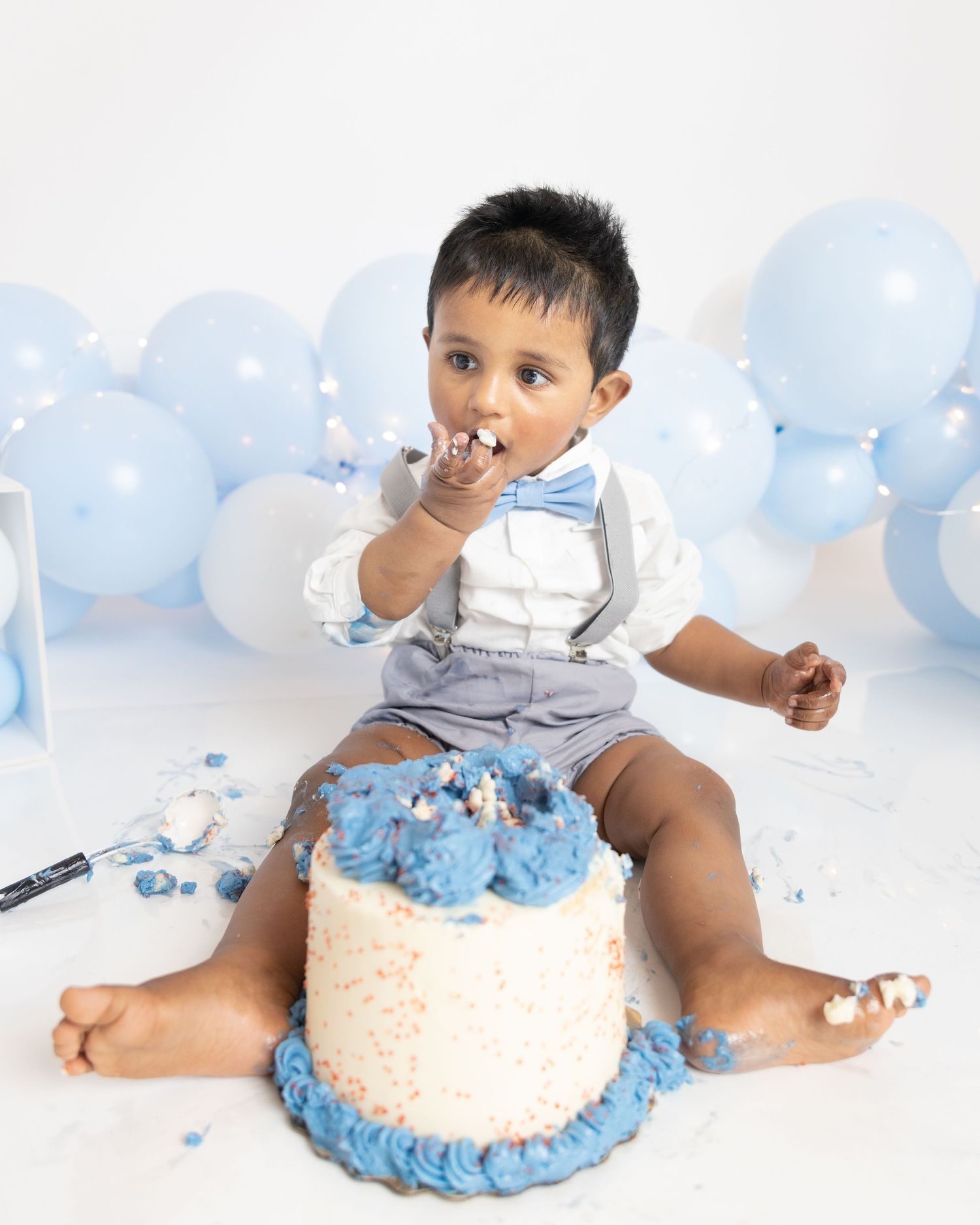 Cake smash photo with baby in blue outfit sitting on white cake with blue frosting.
