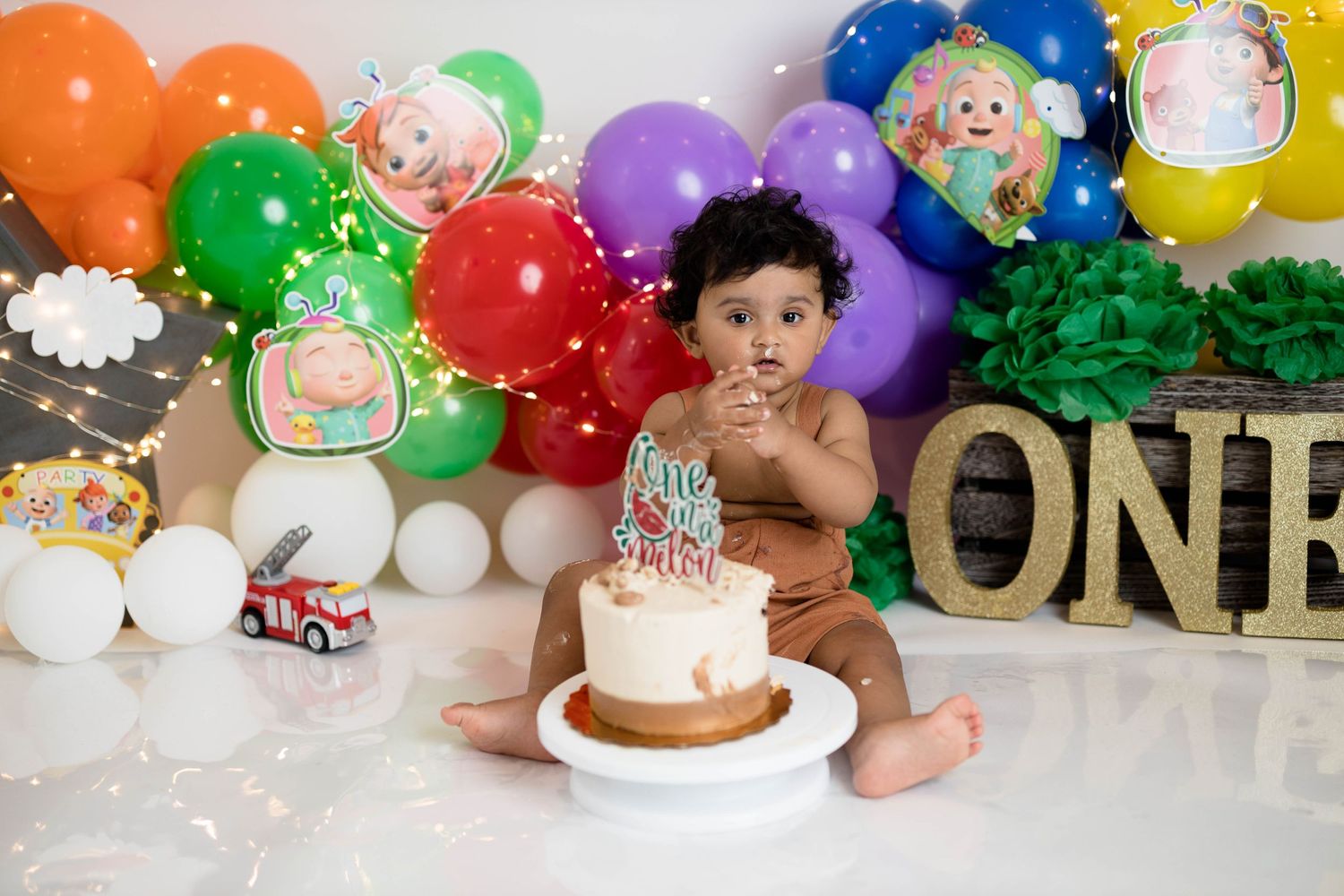 Colorful birthday setup with rainbow balloon arch and gold ONE letters against white backdrop.