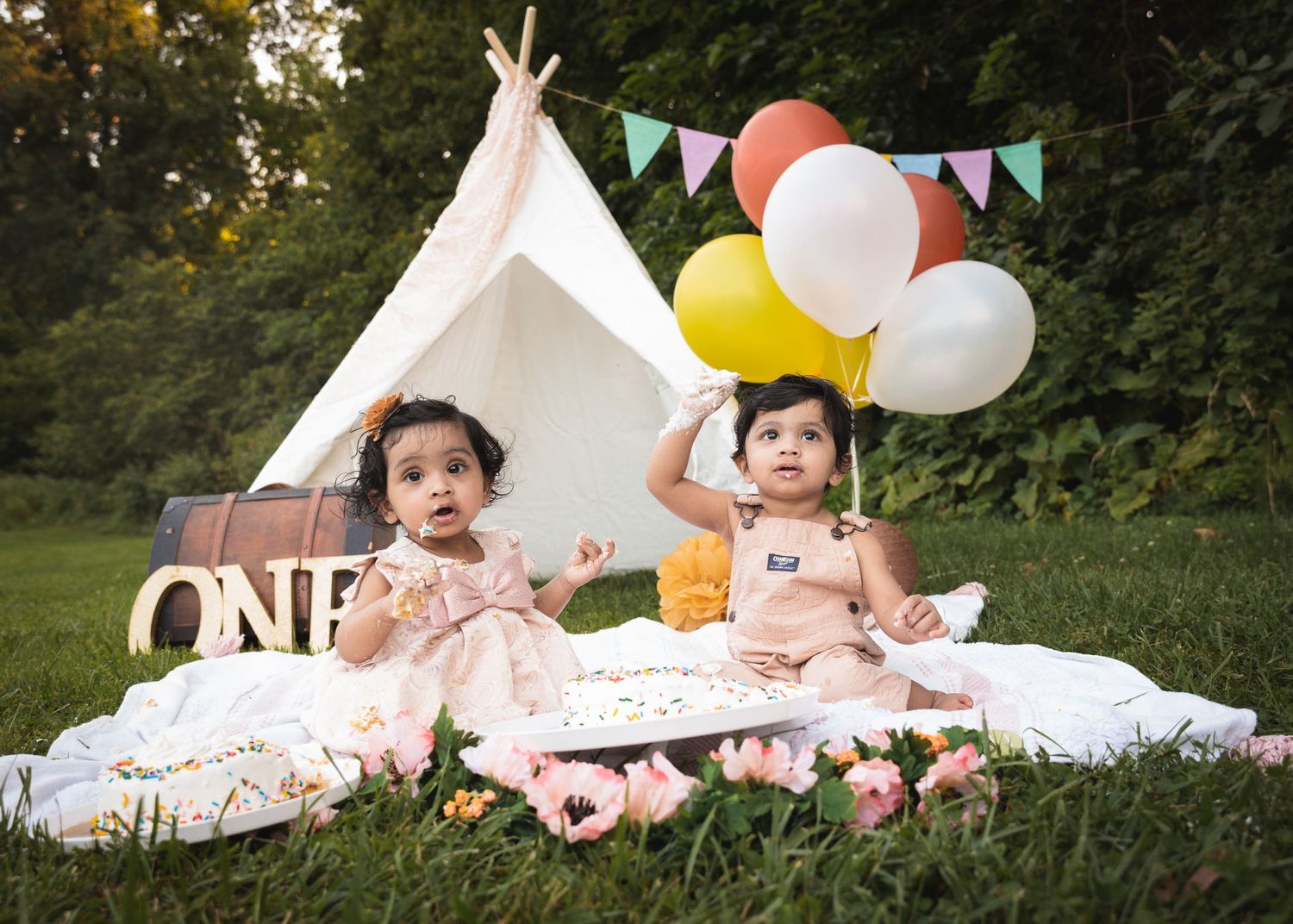 First birthday celebration with teepee, balloons and two babies in party dresses on white blanket.