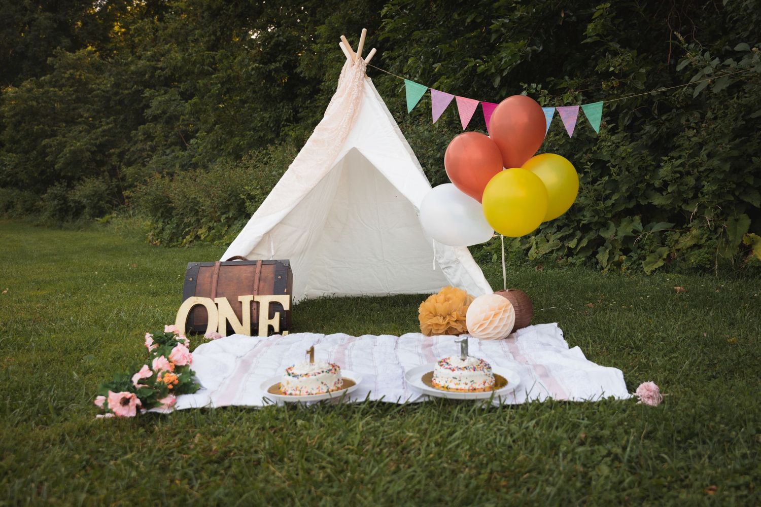 Rustic first birthday setup with white teepee, balloons and bunting against green backdrop.