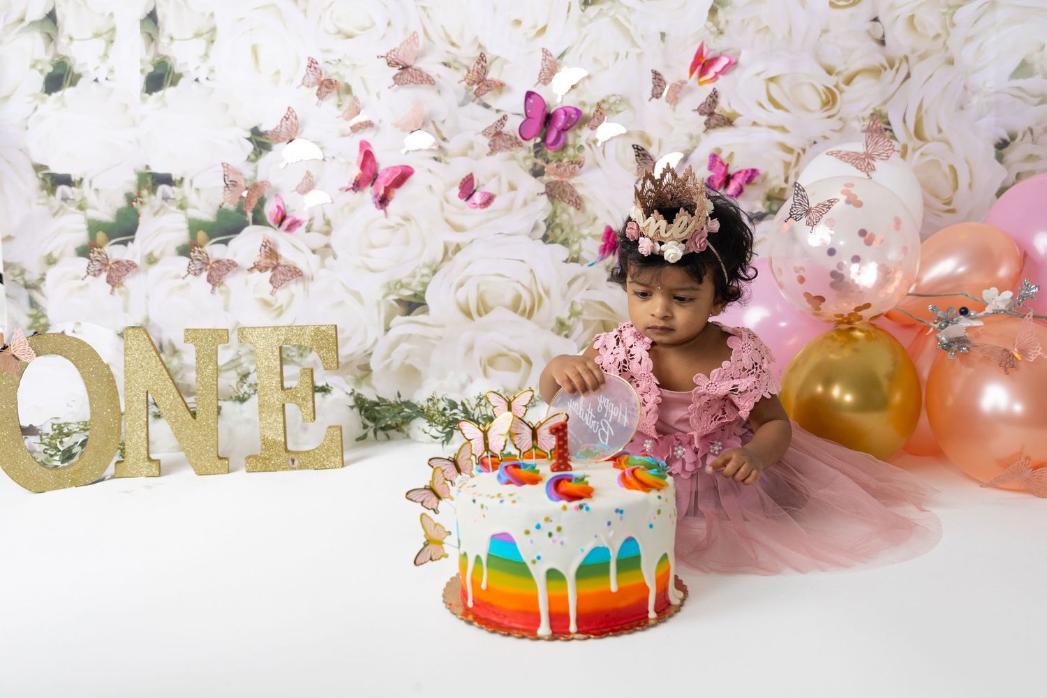 Whimsical first birthday setup with rainbow cake, gold letters, butterflies and pink balloons.