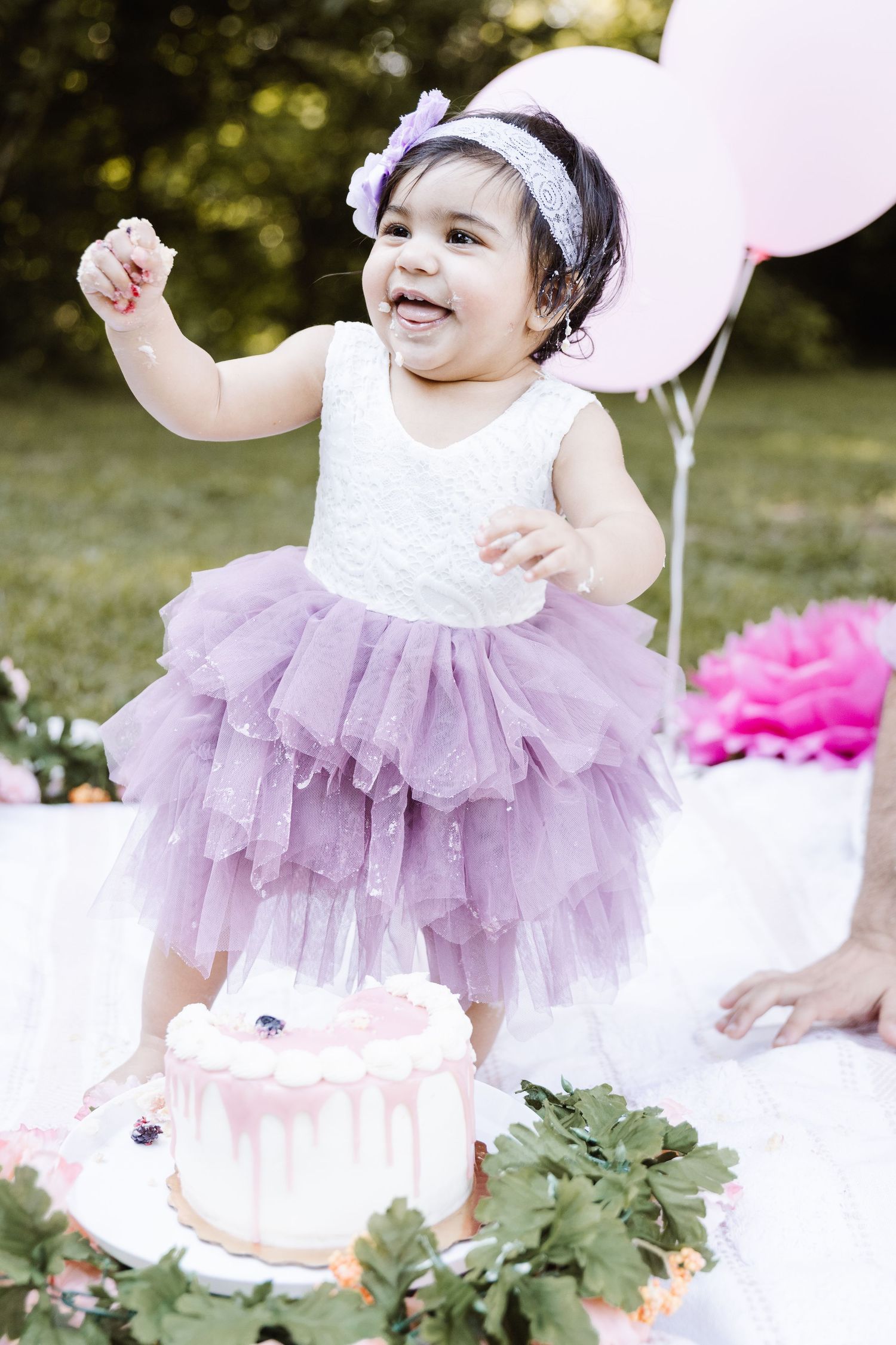 Baby in a lavender tutu dress celebrating first birthday with pink balloons and cake outdoors.