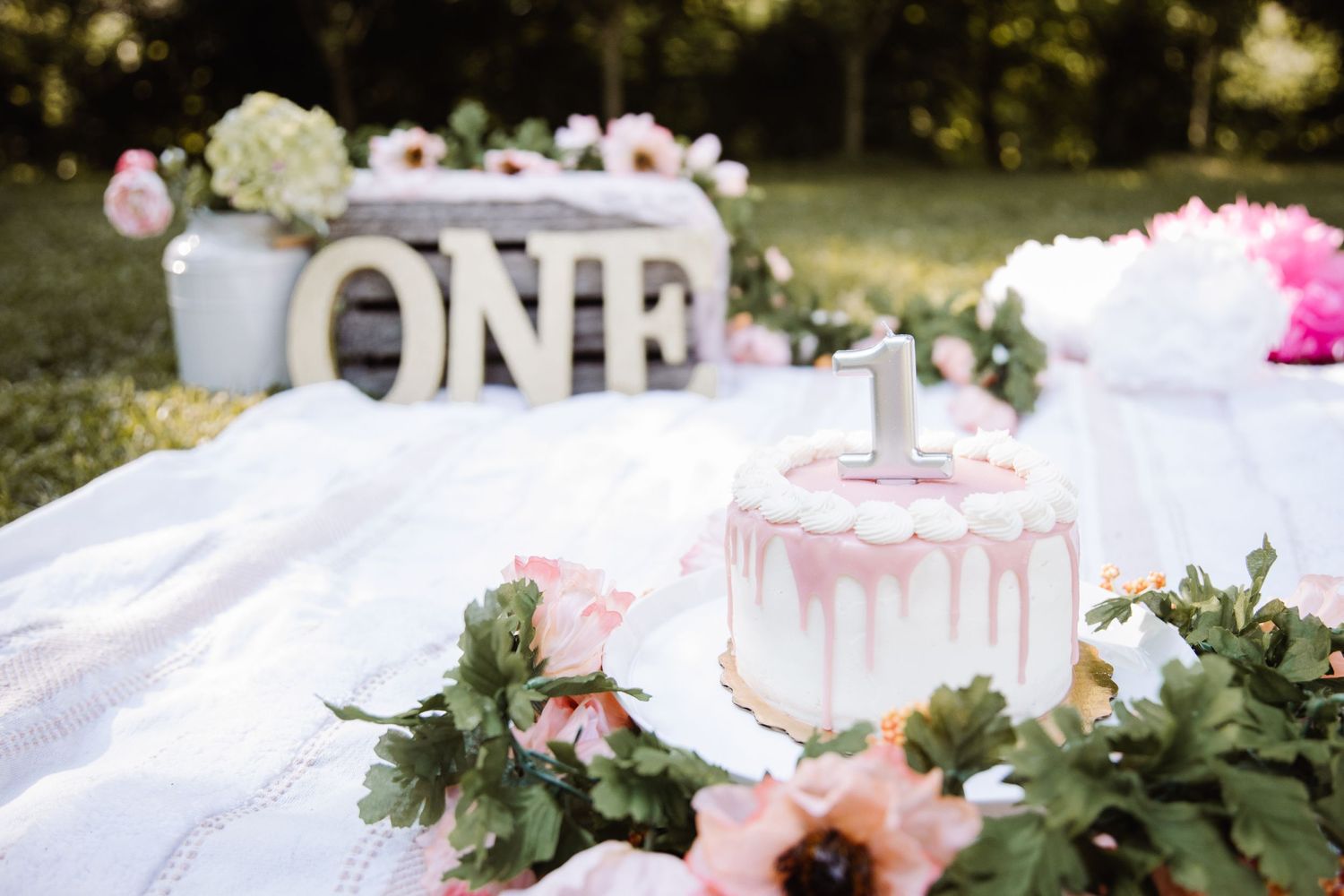 First birthday party setup with 'ONE' sign, pink drip cake, and floral decorations on white blanket.